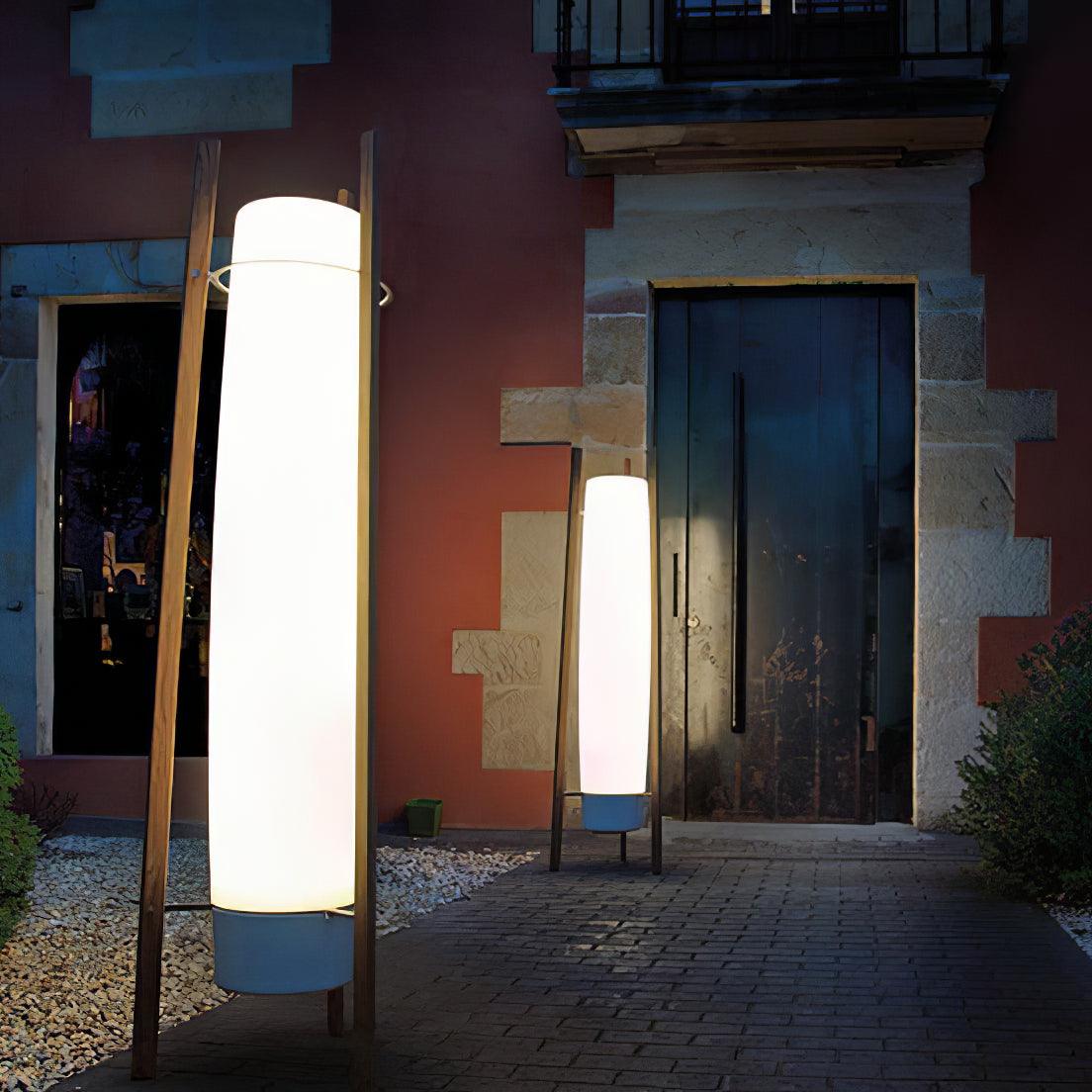 Night courtyard view of two glowing column lamps with wooden supports beside a wood door, pebbles and shrubs