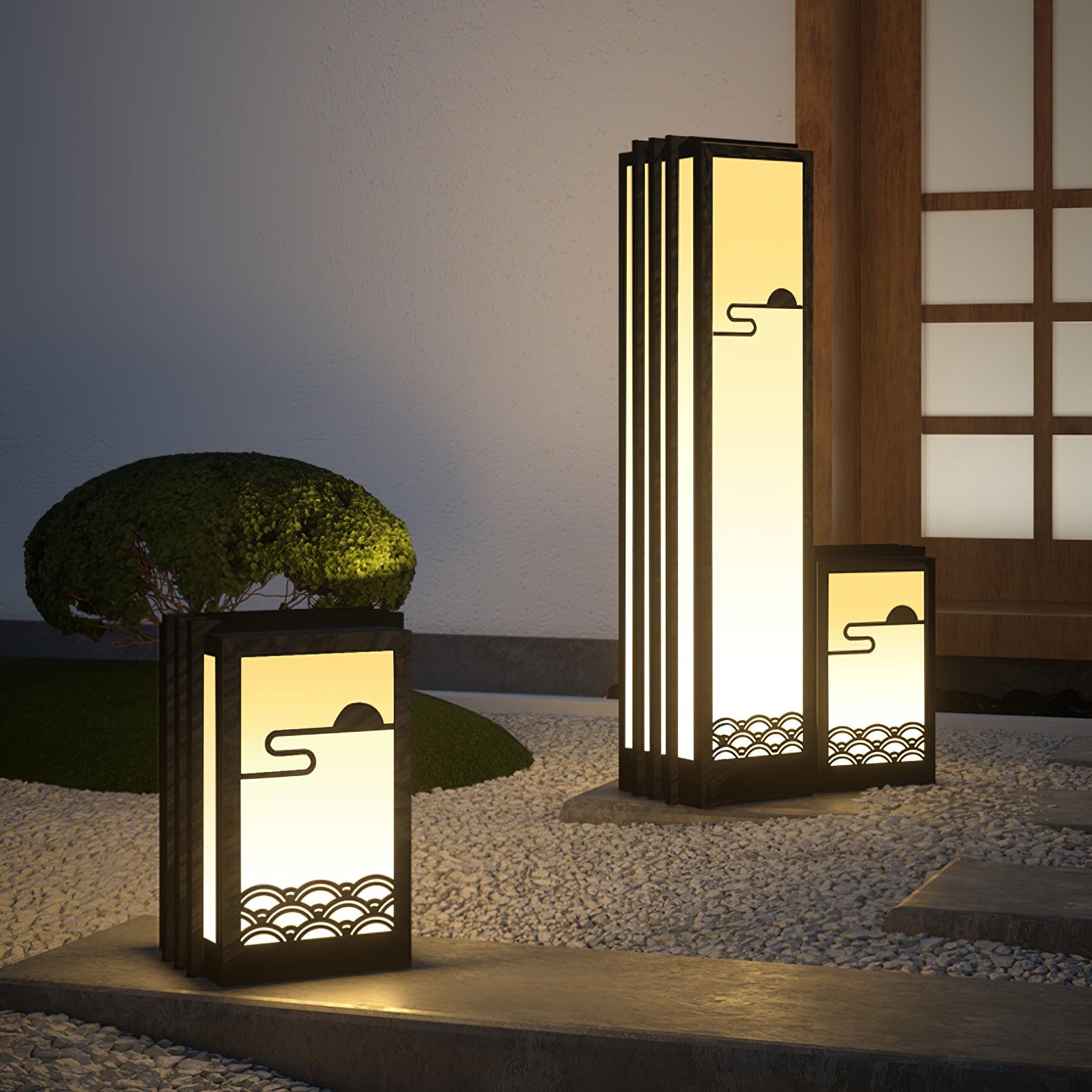Three lit sun-and-wave lanterns by entrance paving, gravel, rounded shrub and wood-grid door