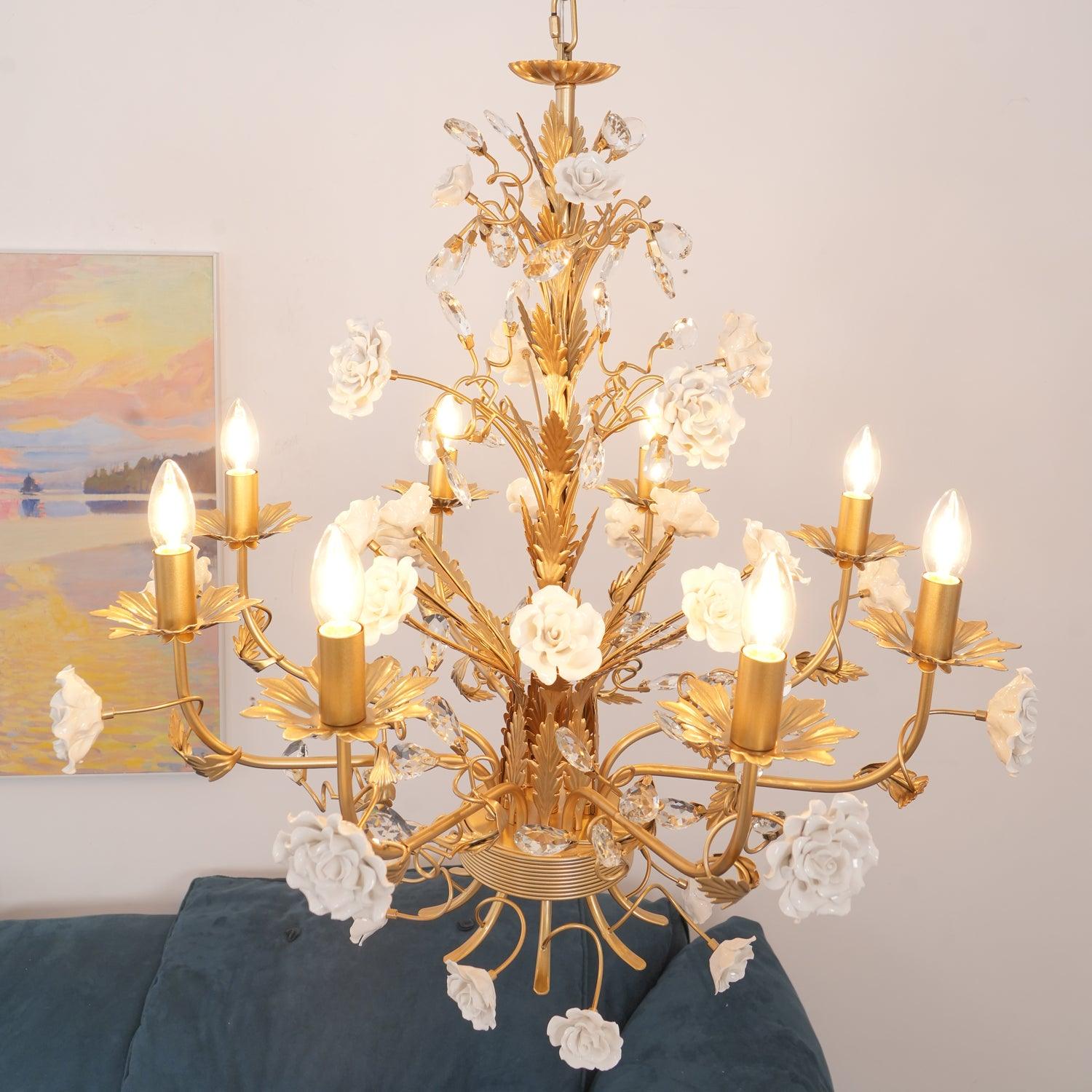 High angled view of lit eight-arm rose chandelier over blue sofa, with white blossoms, gold leaves and clear crystal buds