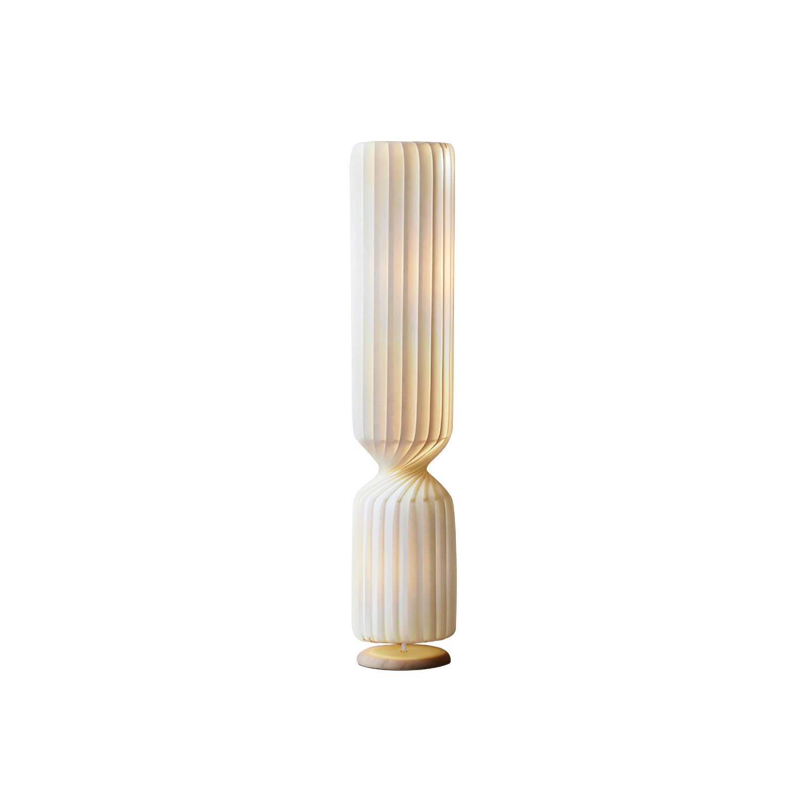 Isolated lit white Twist lamp with elongated pleated upper section, short lower section and pale gold-toned round base