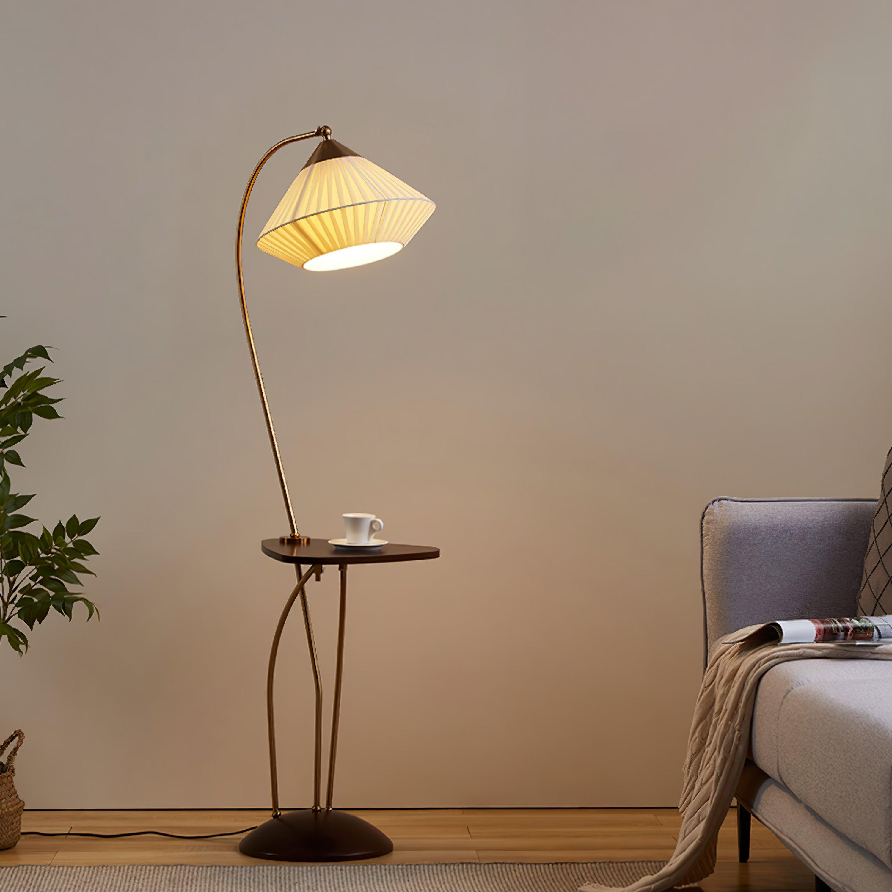 Wide view of Curve’s curved frame, lit beige pleated shade, cup-topped table and dark base beside a grey armchair