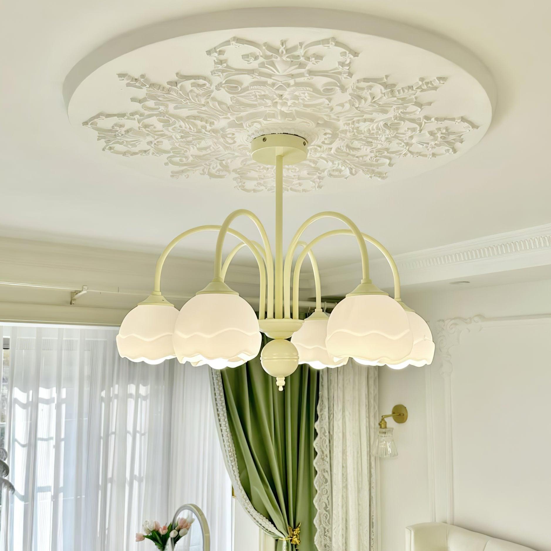 Illuminated cream chandelier with white scalloped shades beneath an ornate ceiling rose beside green curtains