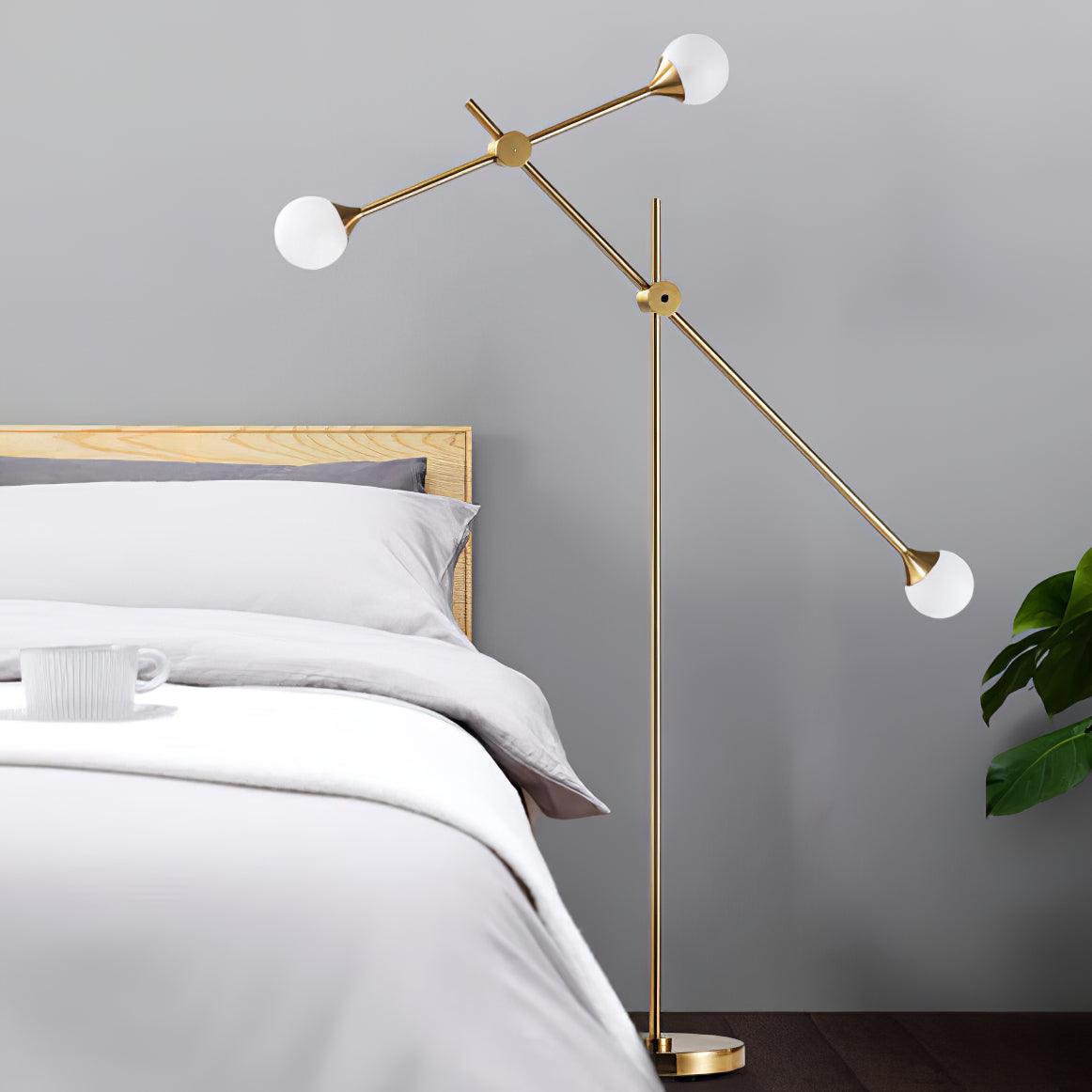 Kemi floor lamp beside a wooden bed frame, with three white globes on angled gold arms against a grey wall