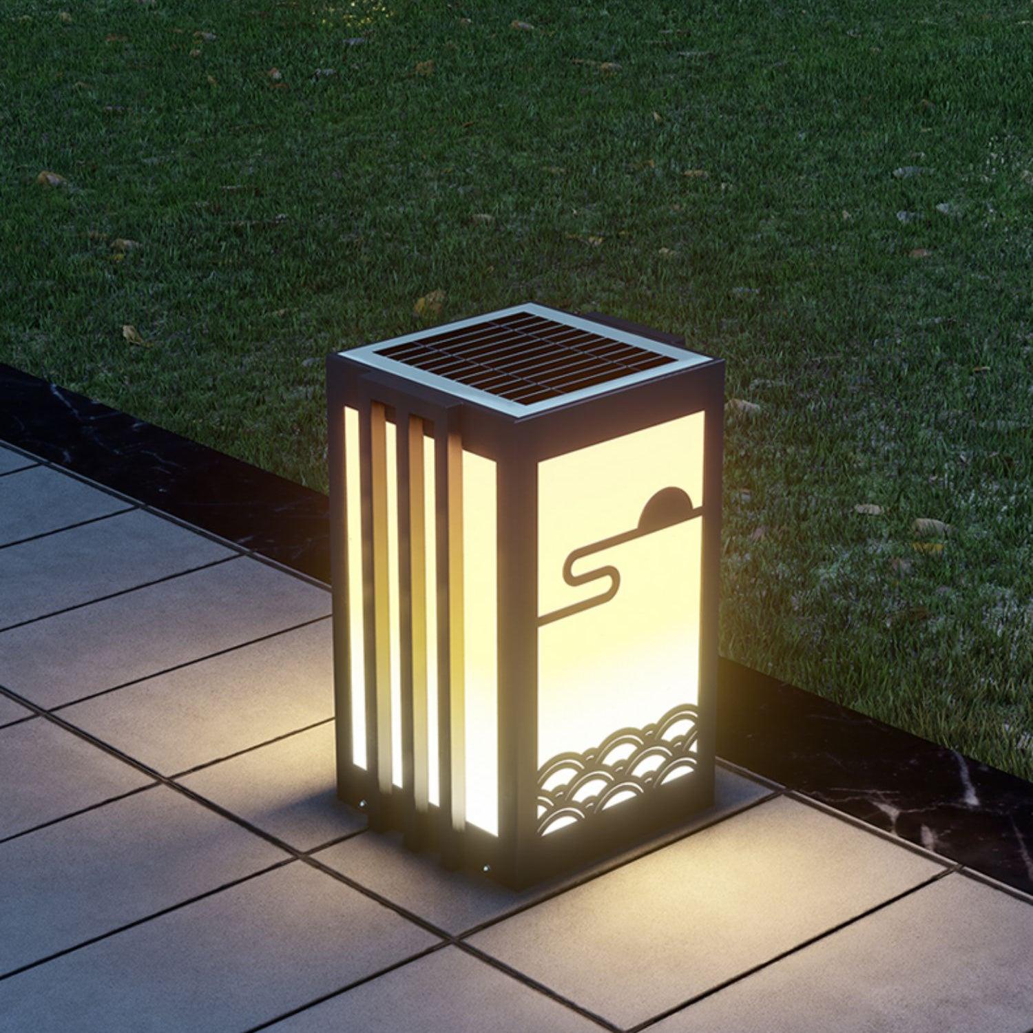 Short solar lantern on tiled garden path beside lawn, showing segmented top panel and illuminated sun-and-wave front