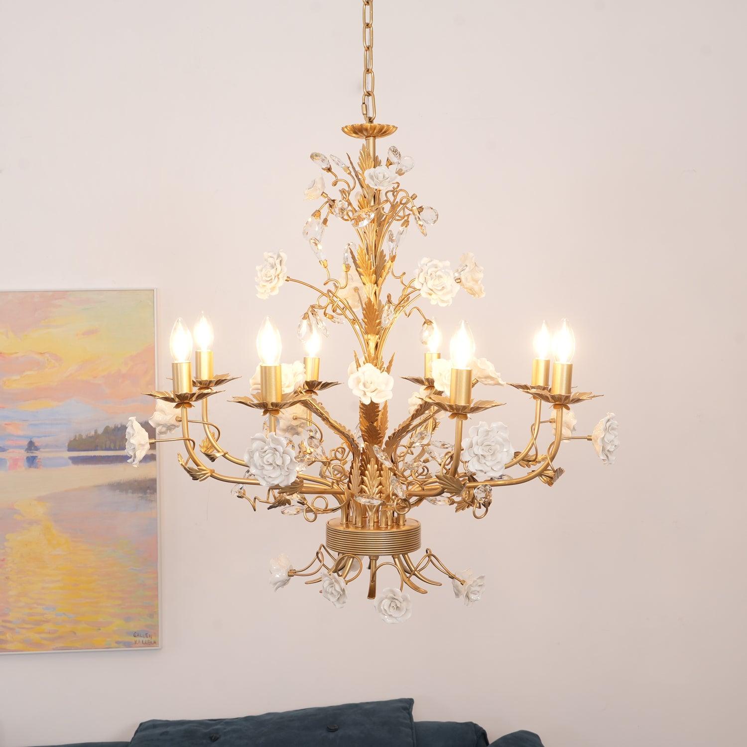 Close broad lit rose chandelier with golden candle arms and clear buds, beside a pastel landscape above blue sofa cushions