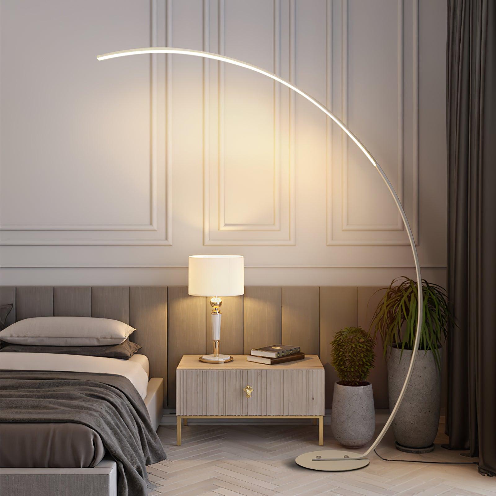 White Arc floor lamp beside bed, nightstand with separate table lamp and books, and tall potted plants near curtain