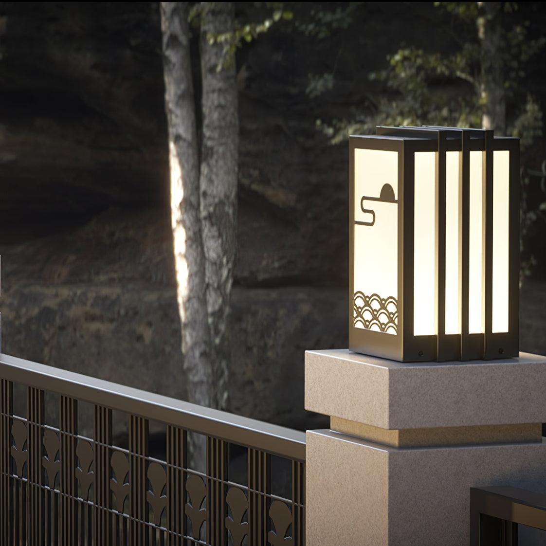 Broad lit sun-and-wave lantern on masonry gatepost, beside dark decorative metal fence and tree