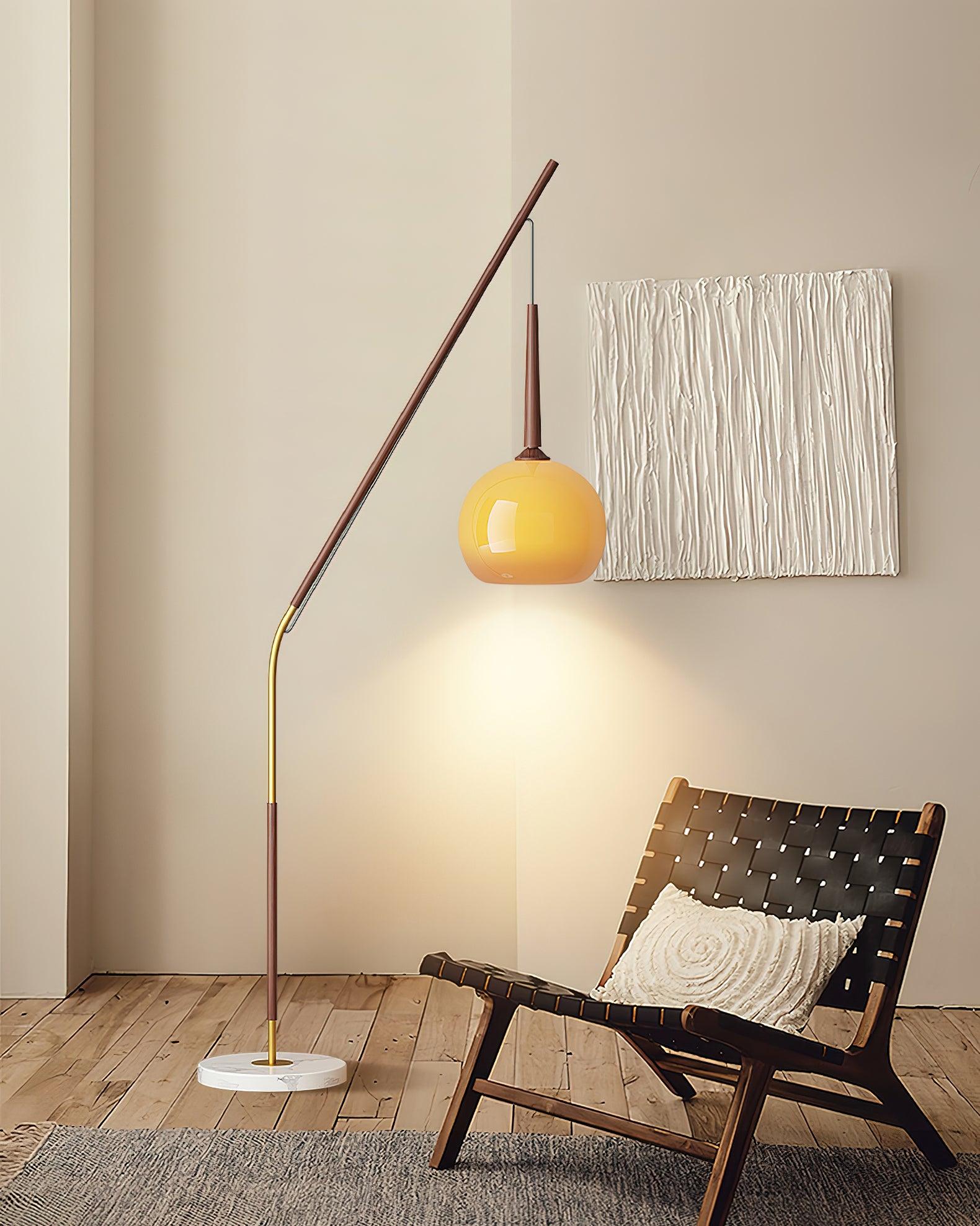 Dark wood-colour angled floor lamp with orange shade and marble base beside a woven-strap chair and textured wall art
