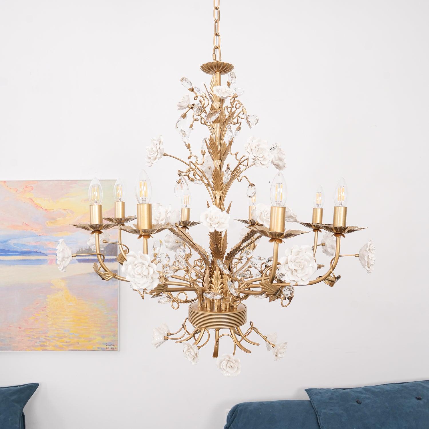 Unlit eight-light floral chandelier beside a pastel landscape above blue sofa cushions, with white roses and clear crystal buds