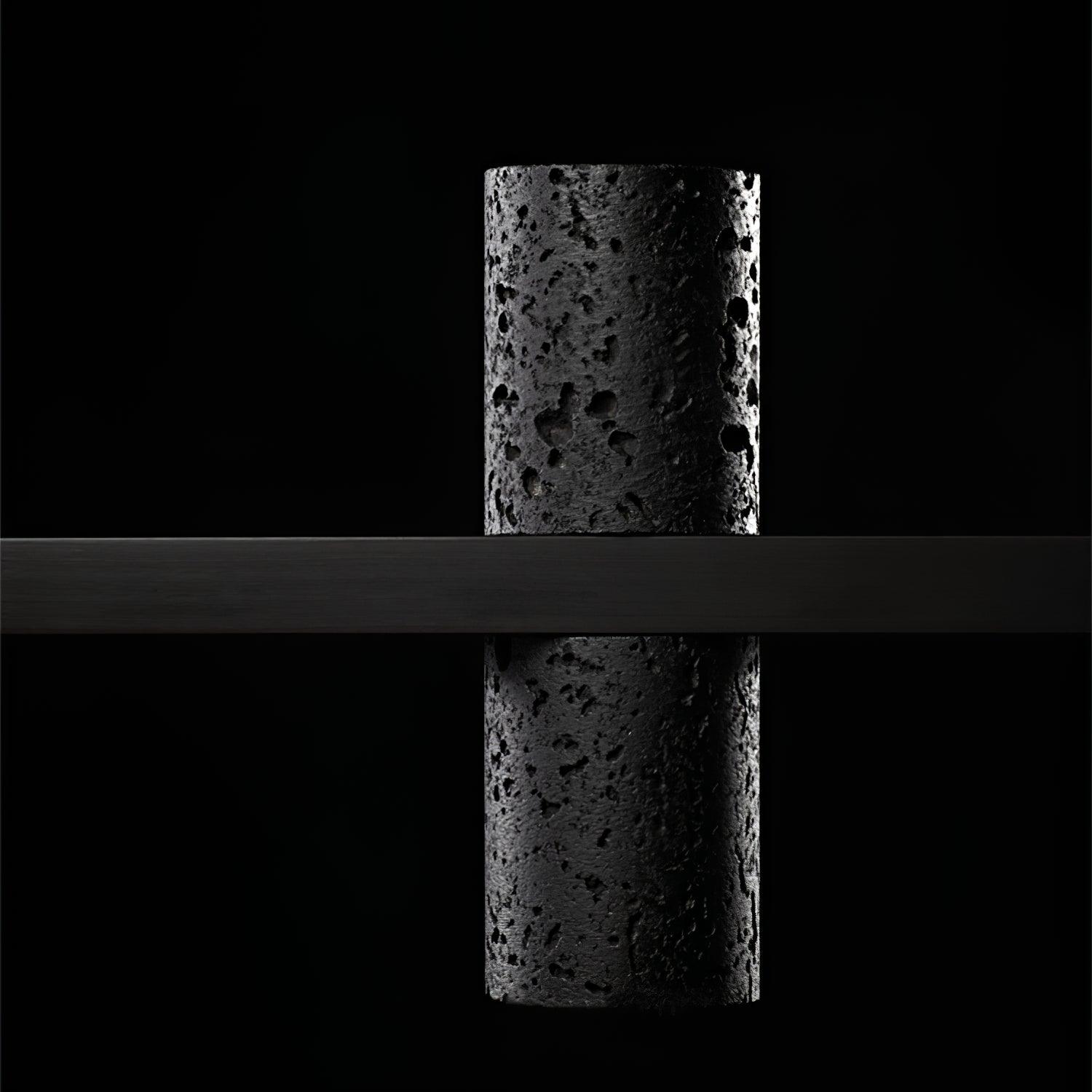 Unlit close side view of one pitted black cylindrical stone element intersected by smooth horizontal black bar.