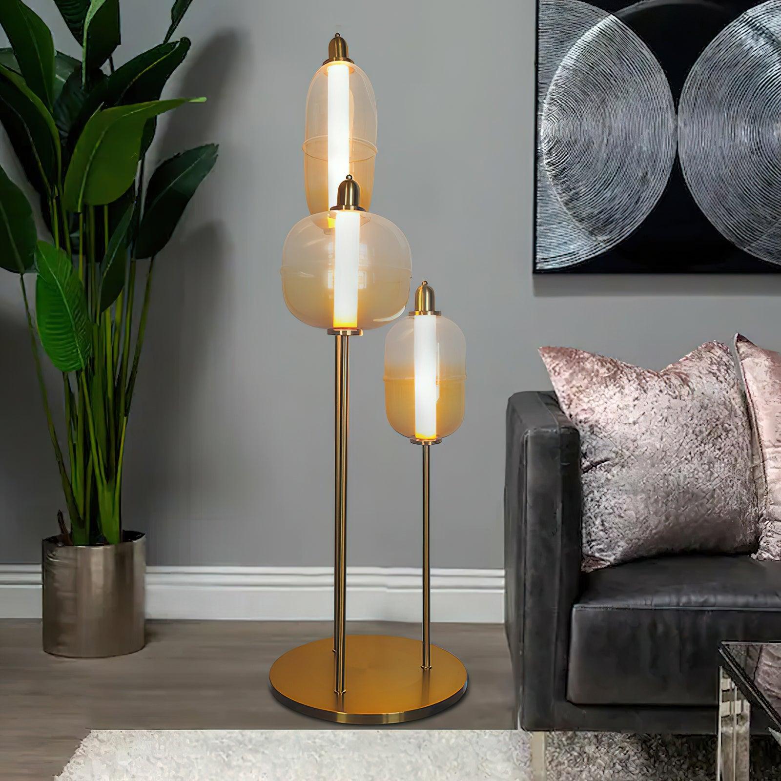 Three amber glass shades on a gold stand beside a dark sofa, pink pillows, tall plant and black-and-white circle print