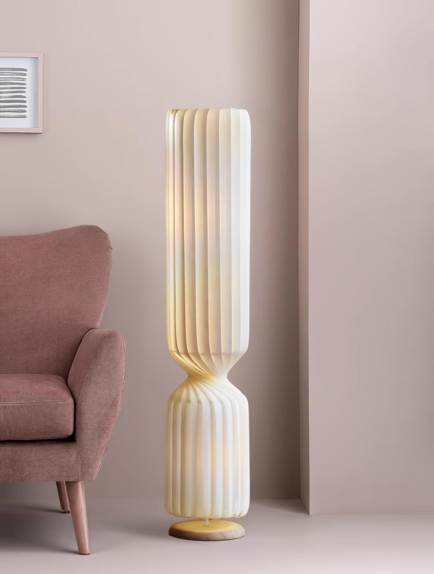 Lit white pleated Twist floor lamp beside pink upholstered chair and pink wall, with warm-toned folds and pale round base