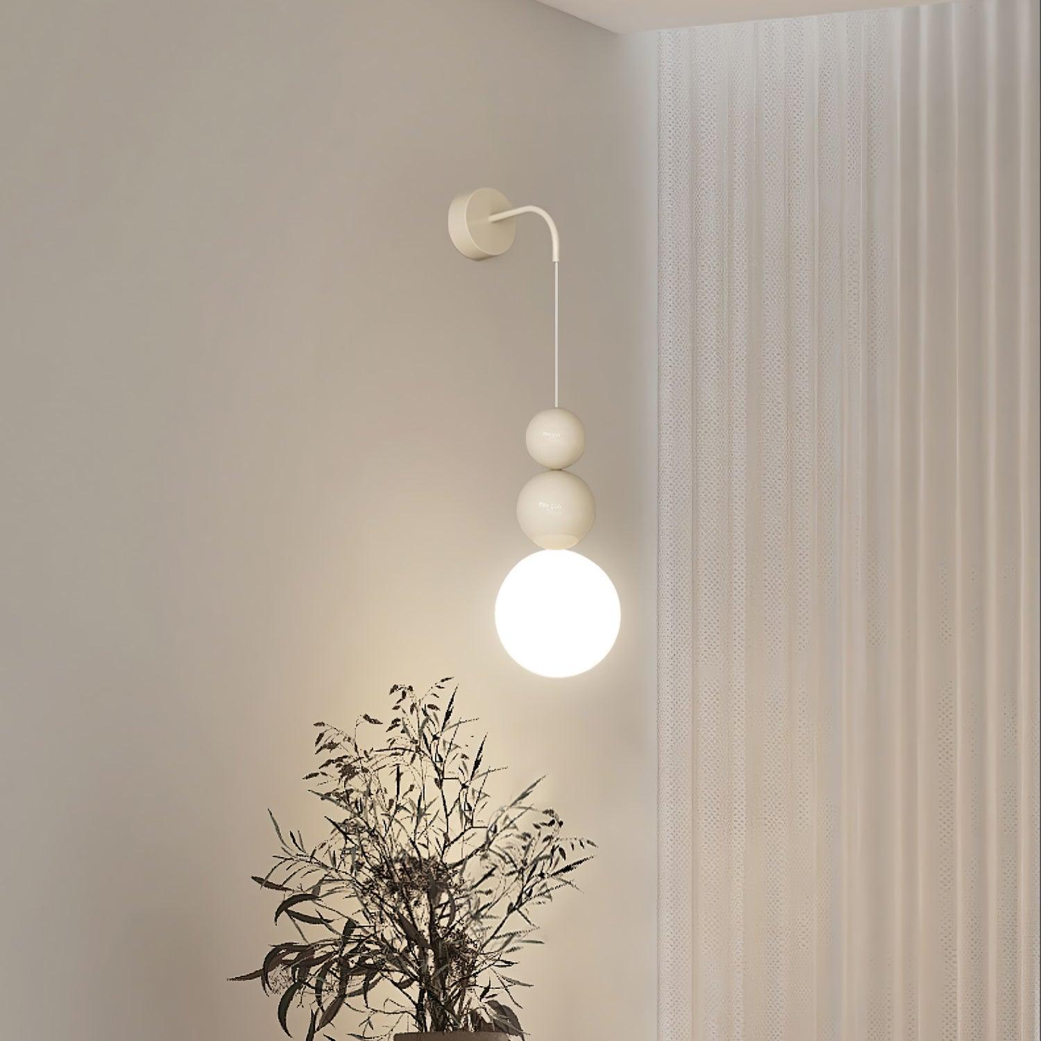 Lit beige Three Ball wall light beside a window curtain and plant