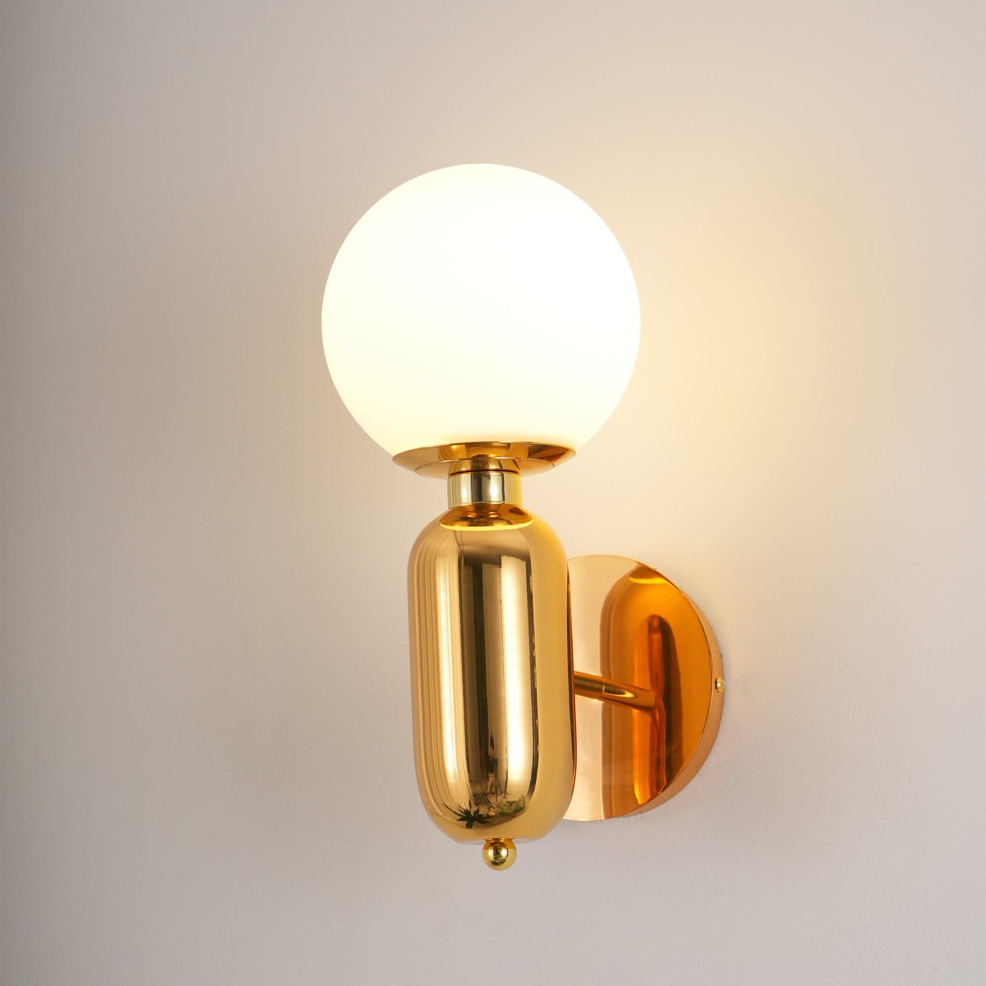 Angled gold-toned Aballs sconce lit on pale wall, white globe, reflective body, round plate and short projecting arm