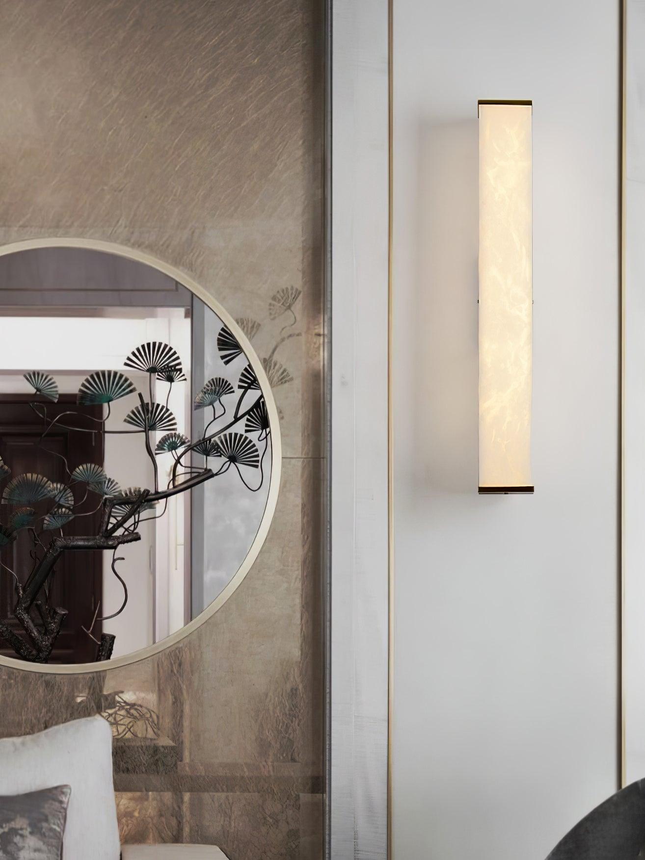 Tall view of an illuminated rectangular alabaster wall light beside a round mirror and pale vertical wall trim