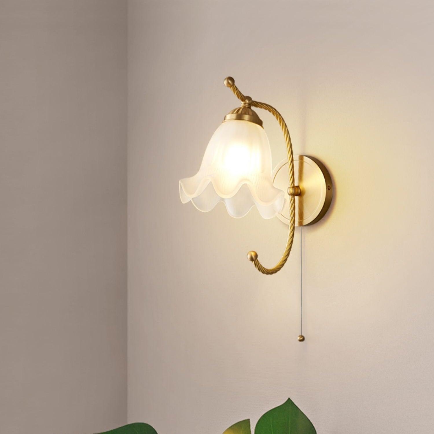 Lit floral glass wall lamp on plain wall near corner, with curling brass-finish arm, thin pull cord and leaves at lower edge