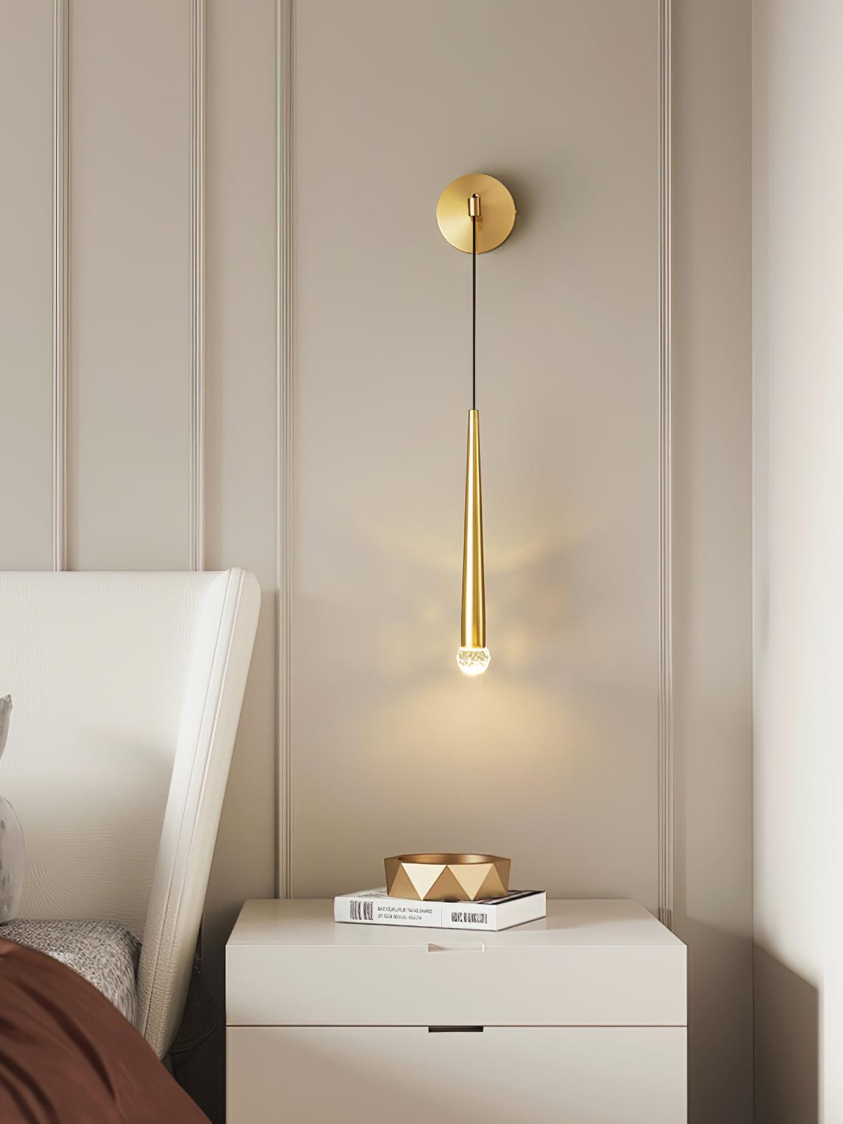 Wider bedside view of a brass-tone crystal-tipped wall light above white drawers, books and a geometric bowl