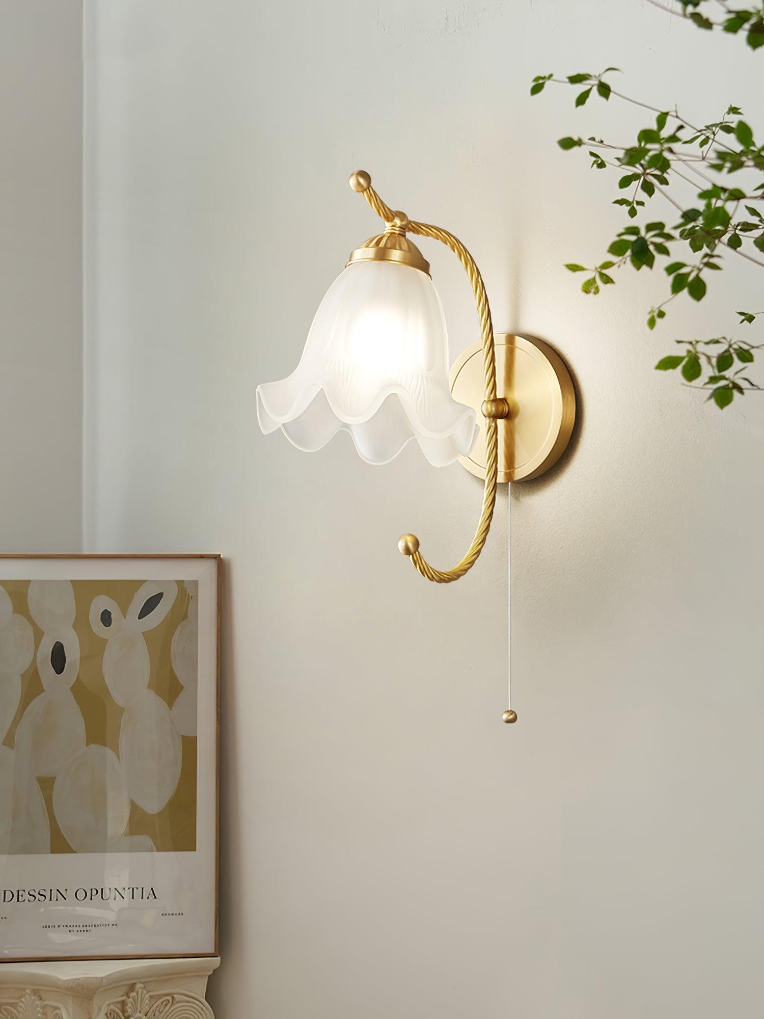 Lit floral glass wall lamp beside framed botanical artwork and leafy branch, with brass-finish plate at right and hanging pull cord