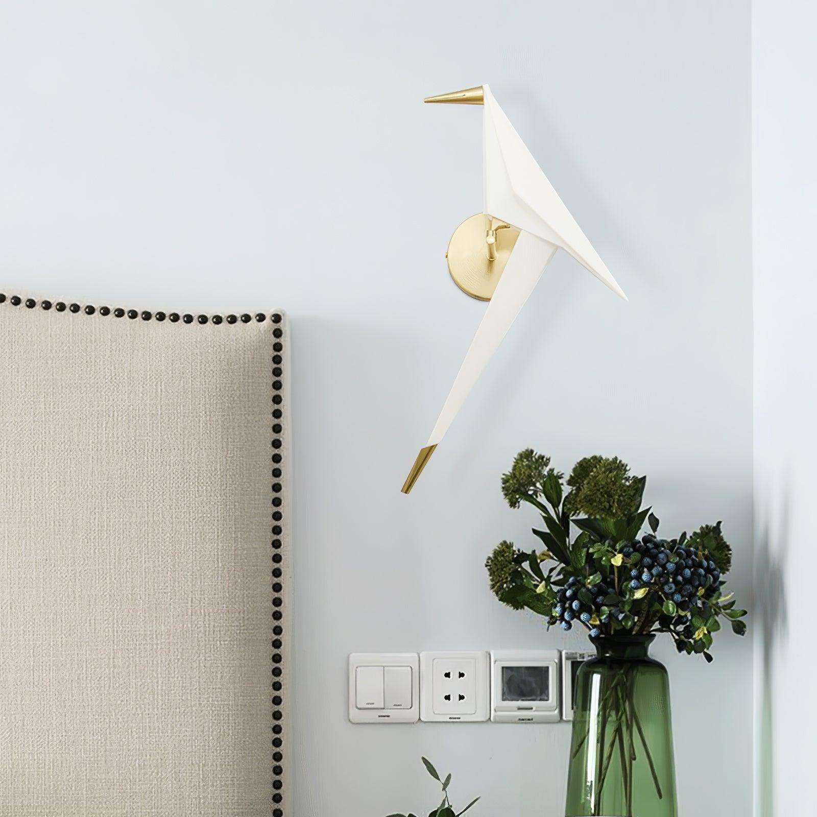 Unlit-looking Gold and White bird wall light beside an upholstered headboard, with wall controls and a green vase of foliage below