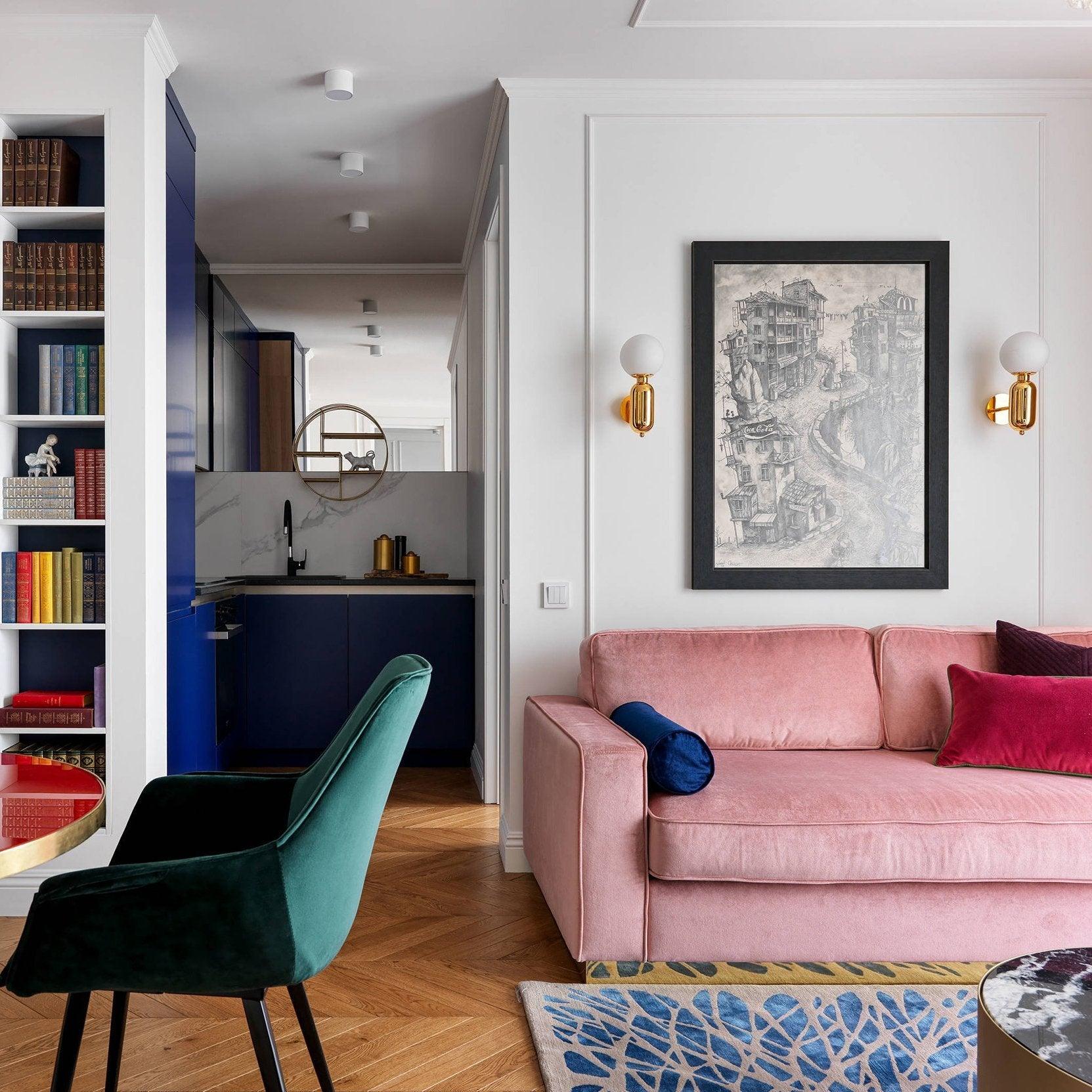 Two gold-toned globe sconces beside framed city drawing above pink sofa, green chair, blue kitchen and bookcase