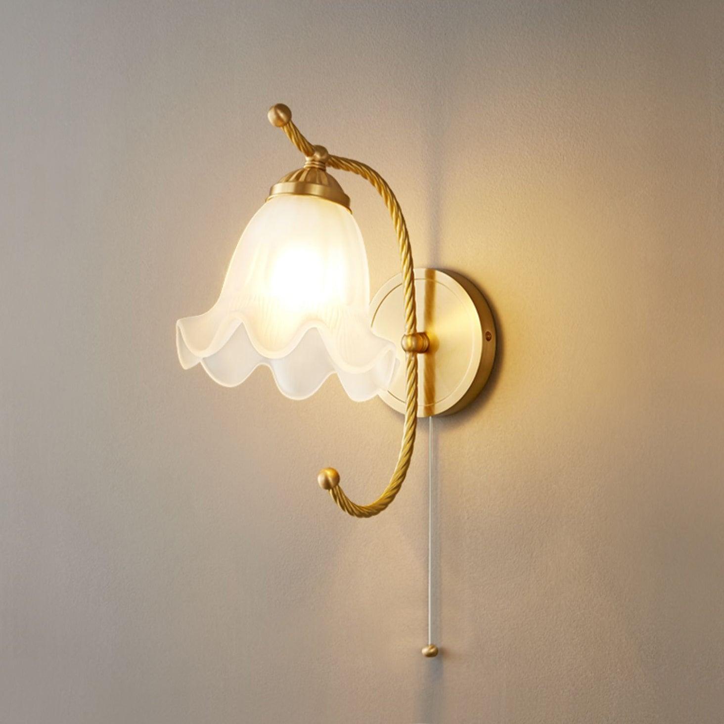 Warm-looking lit scalloped glass lamp against grey wall, with brass-finish curled arm and thin pull cord beneath the plate
