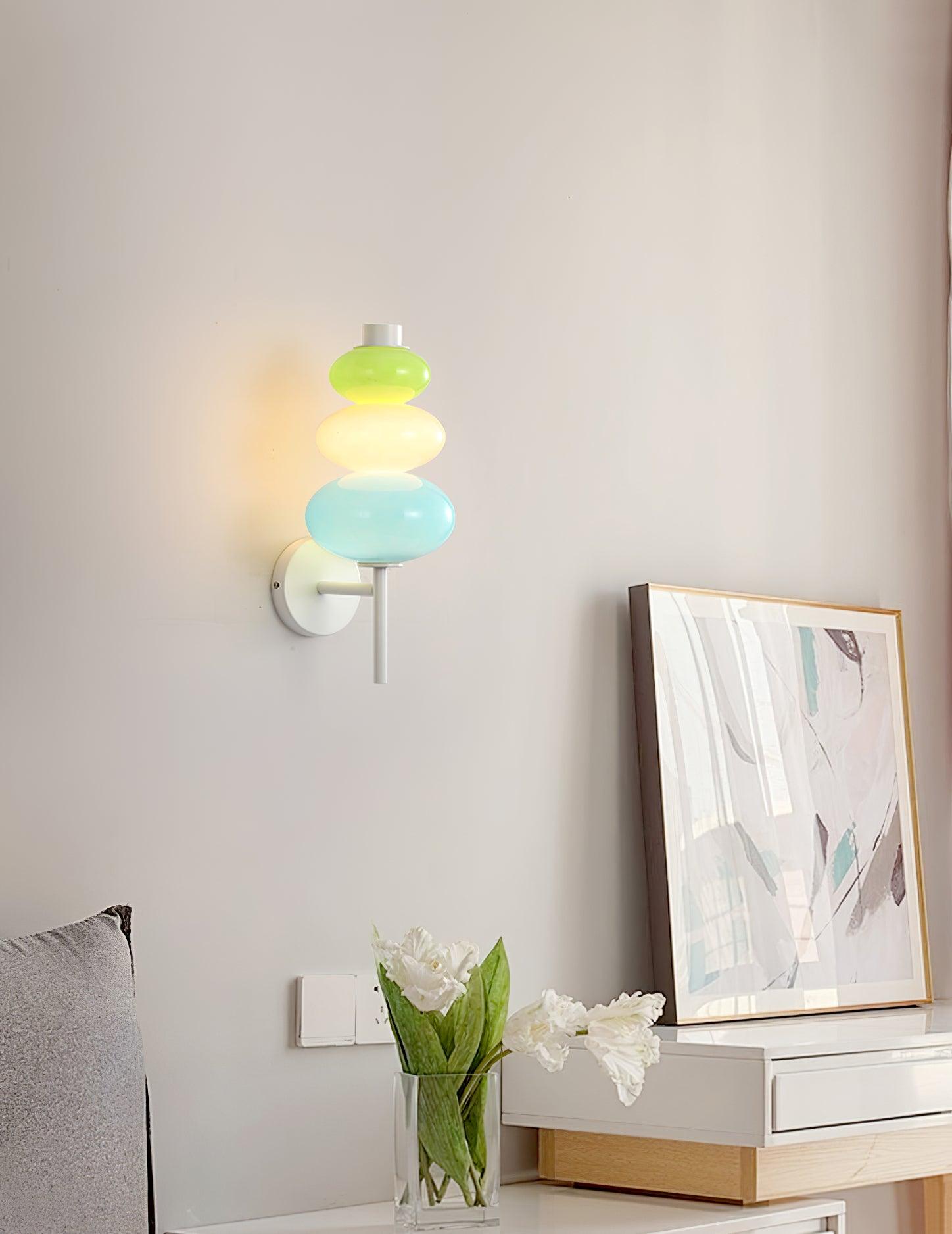 Multicolored glass wall light above a white cabinet near framed pastel artwork and flowers