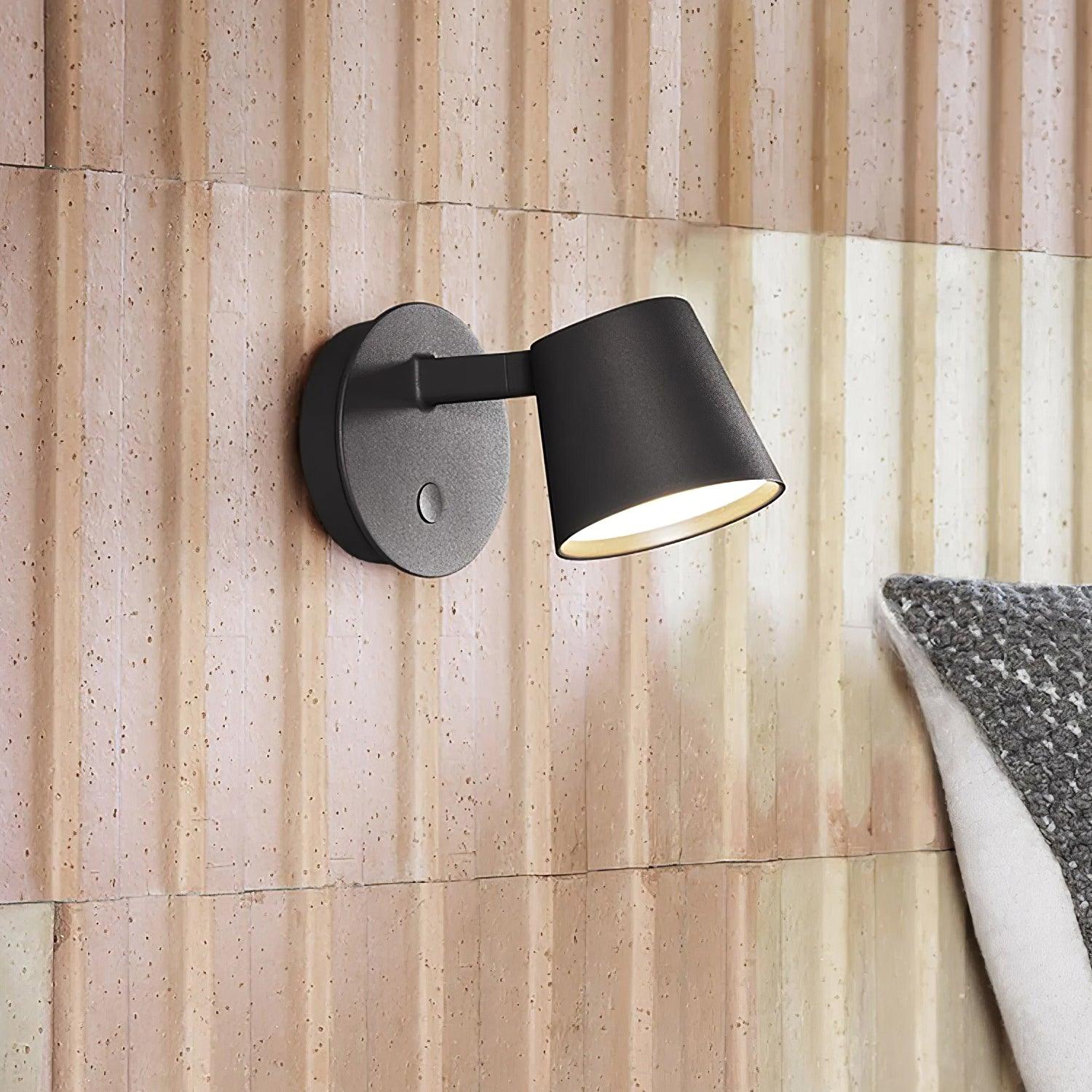 Close black Tip light on pale textured stone wall beside a grey-and-white cushion, with warm-looking lower glow