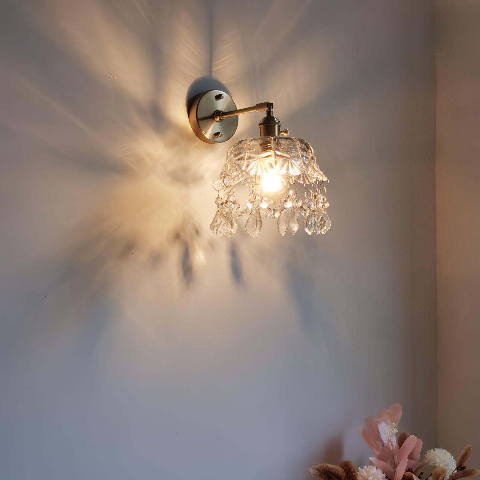 Angled lit crystal-fringed wall light casting patterns on a pale wall, with flowers at the lower corner