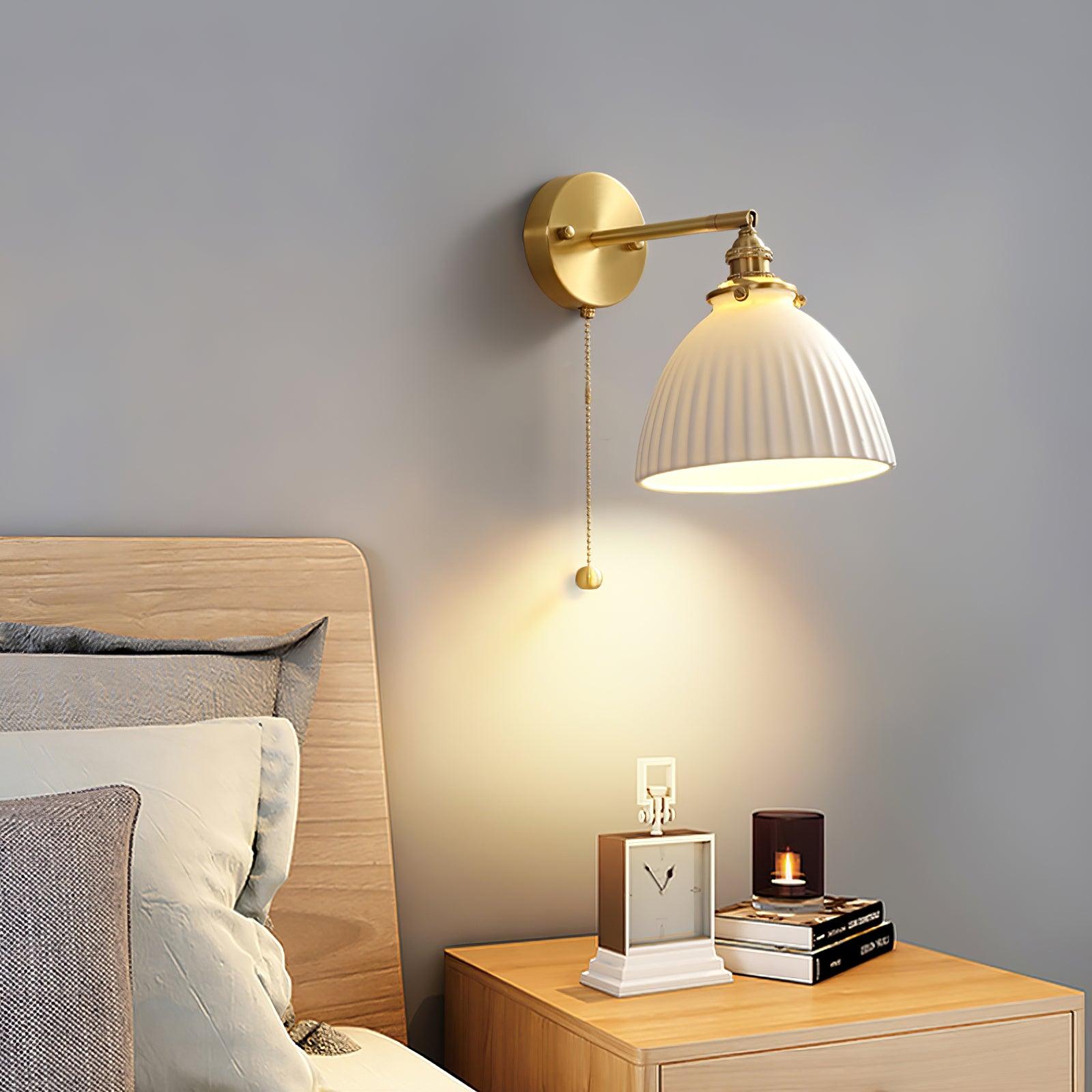 White pleated bedside wall light above a wooden table with clock, books and candle beside a wood headboard