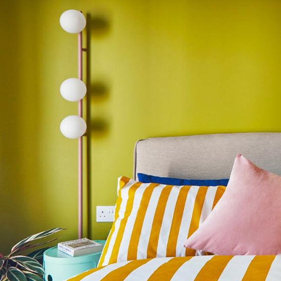 Pink three-globe wall light beside gray bedhead on yellow-green wall, striped cushion, pink pillow and mint bedside table