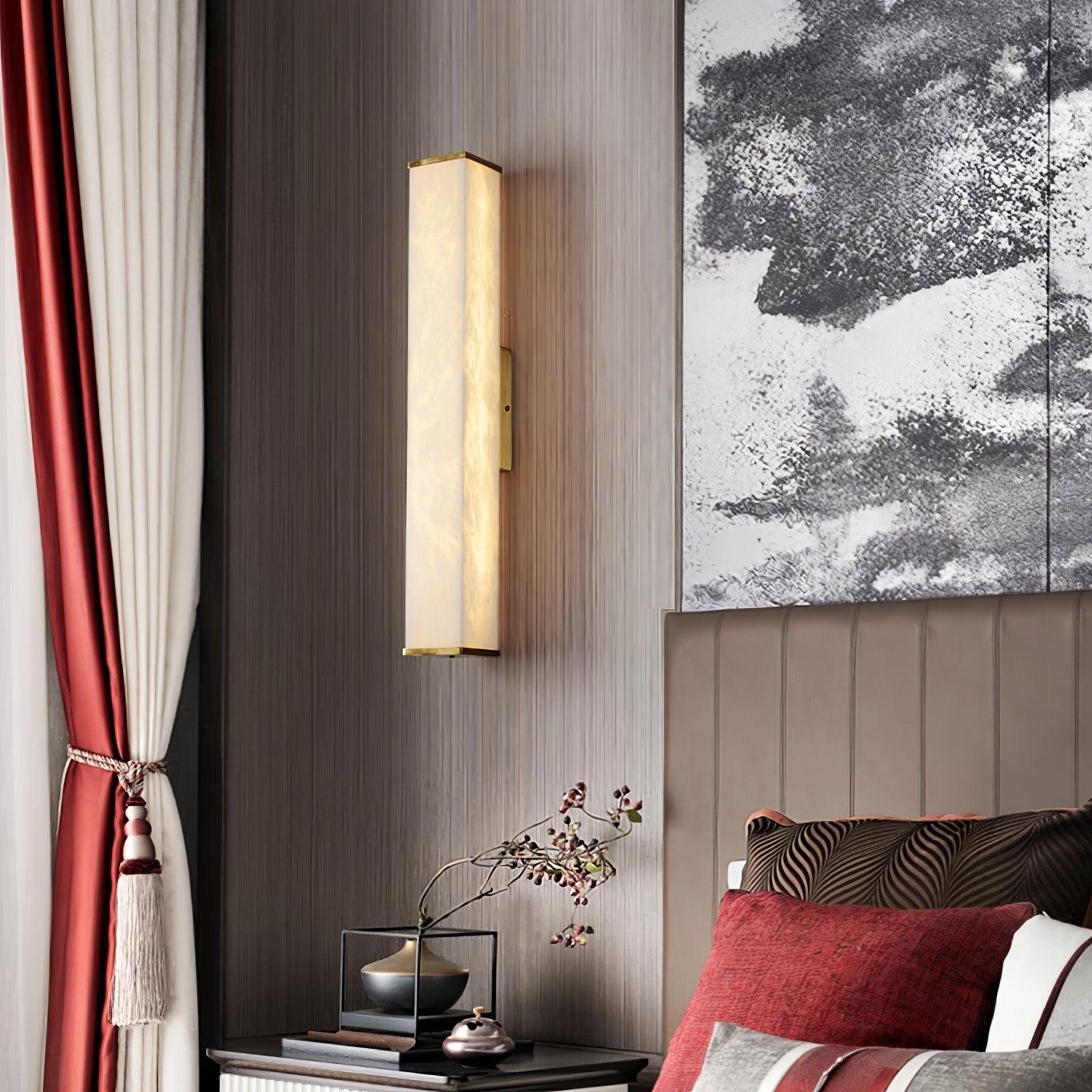 Illuminated alabaster wall light beside a padded headboard, red and cream curtains, and a bedside table with books and a vase