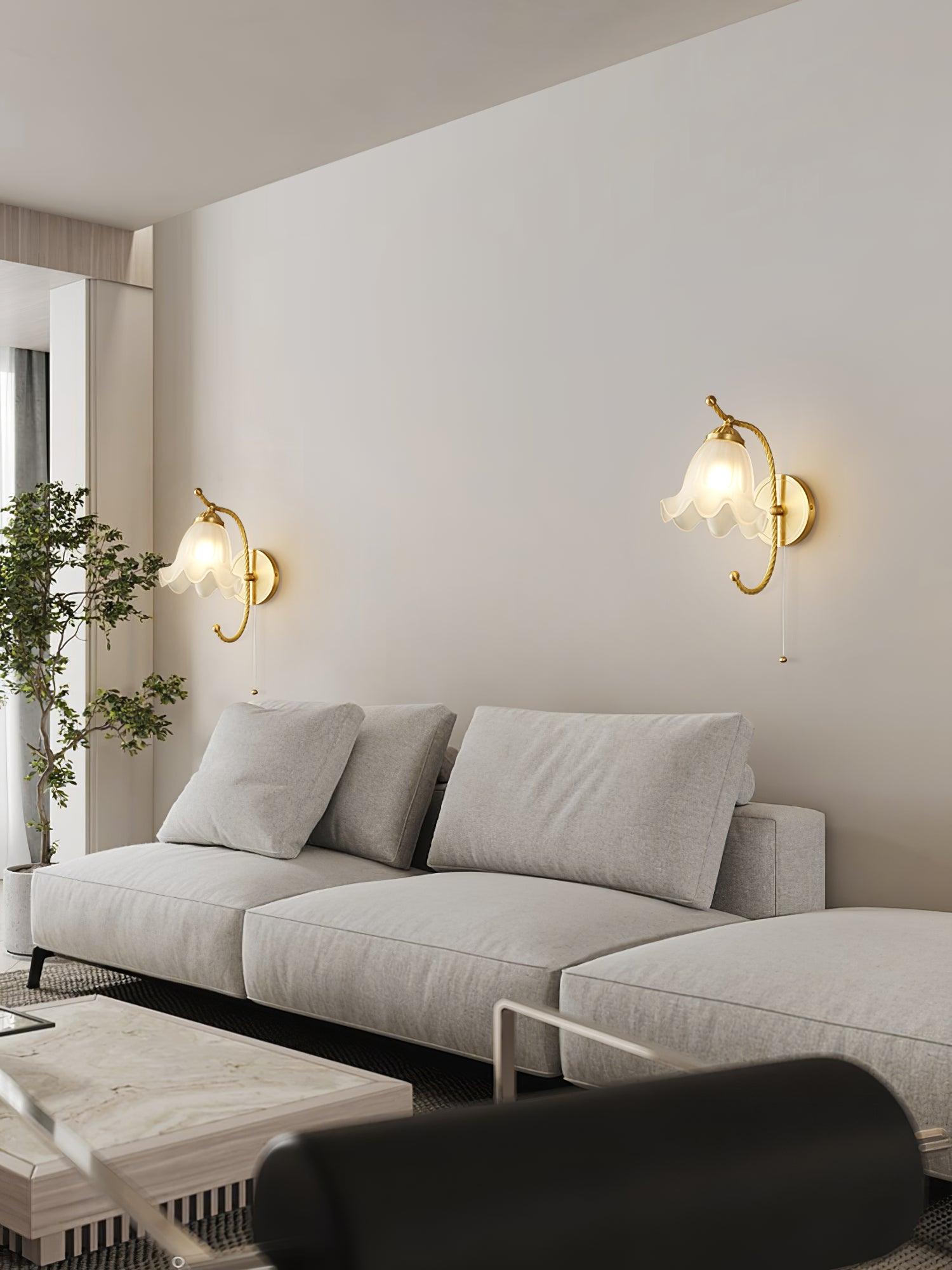 Two matching lit brass gooseneck wall lamps above grey sofa, beside indoor tree and marble-look coffee table