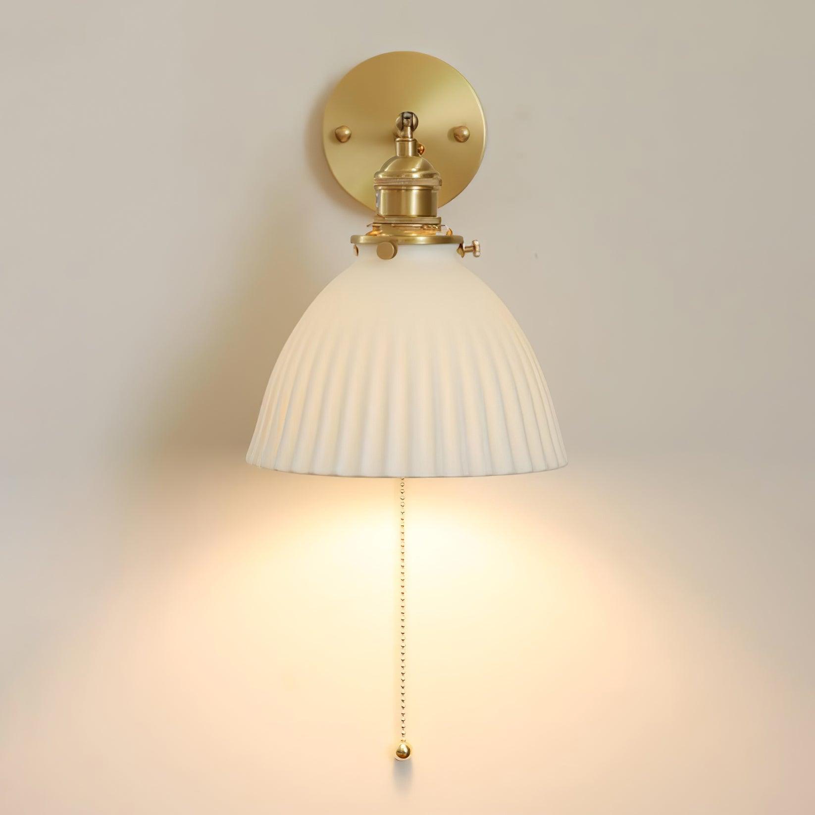 Near-frontal white pleated wall light casting light below the shade, with round brass plate and bead chain