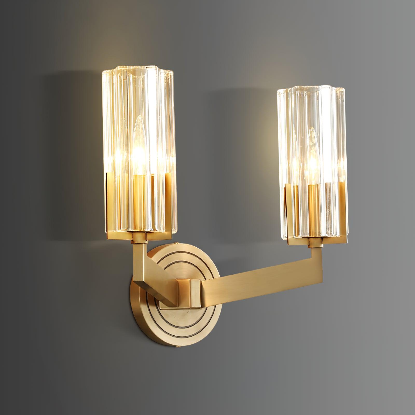 Oblique lit twin-shade Kayla on grey wall with nearer left glass cylinder, angular brass arms and concentric-ring plate