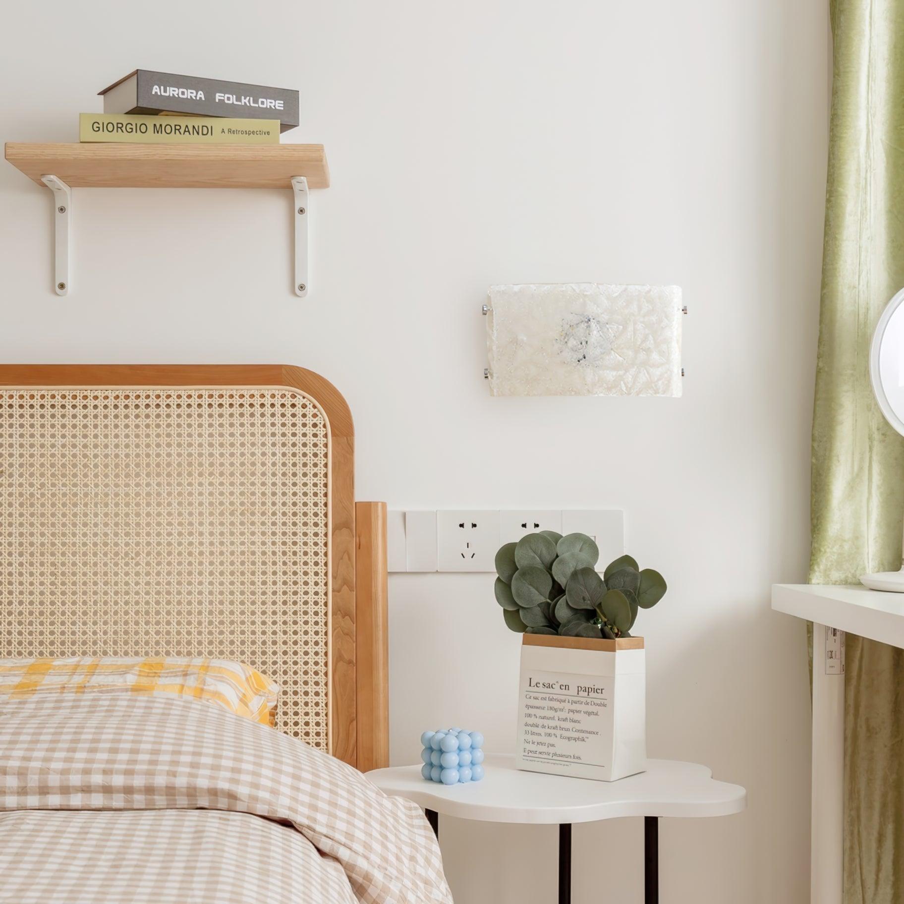 Textured white glass wall light beside a cane headboard, books, green curtain and a blue bubble-shaped candle