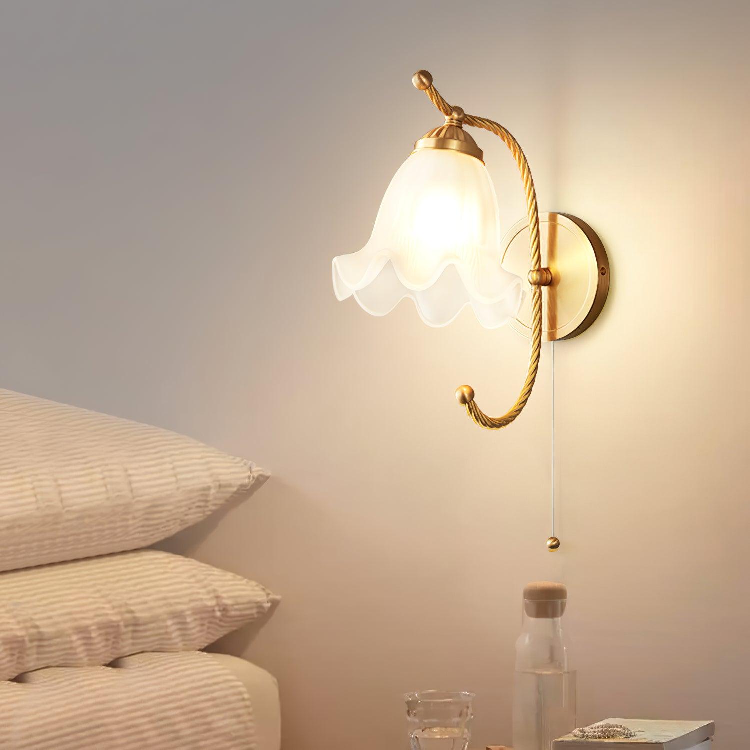 Lit brass gooseneck lamp beside stacked pale pillows and water bottle, with white scalloped shade and pull cord above bedside table