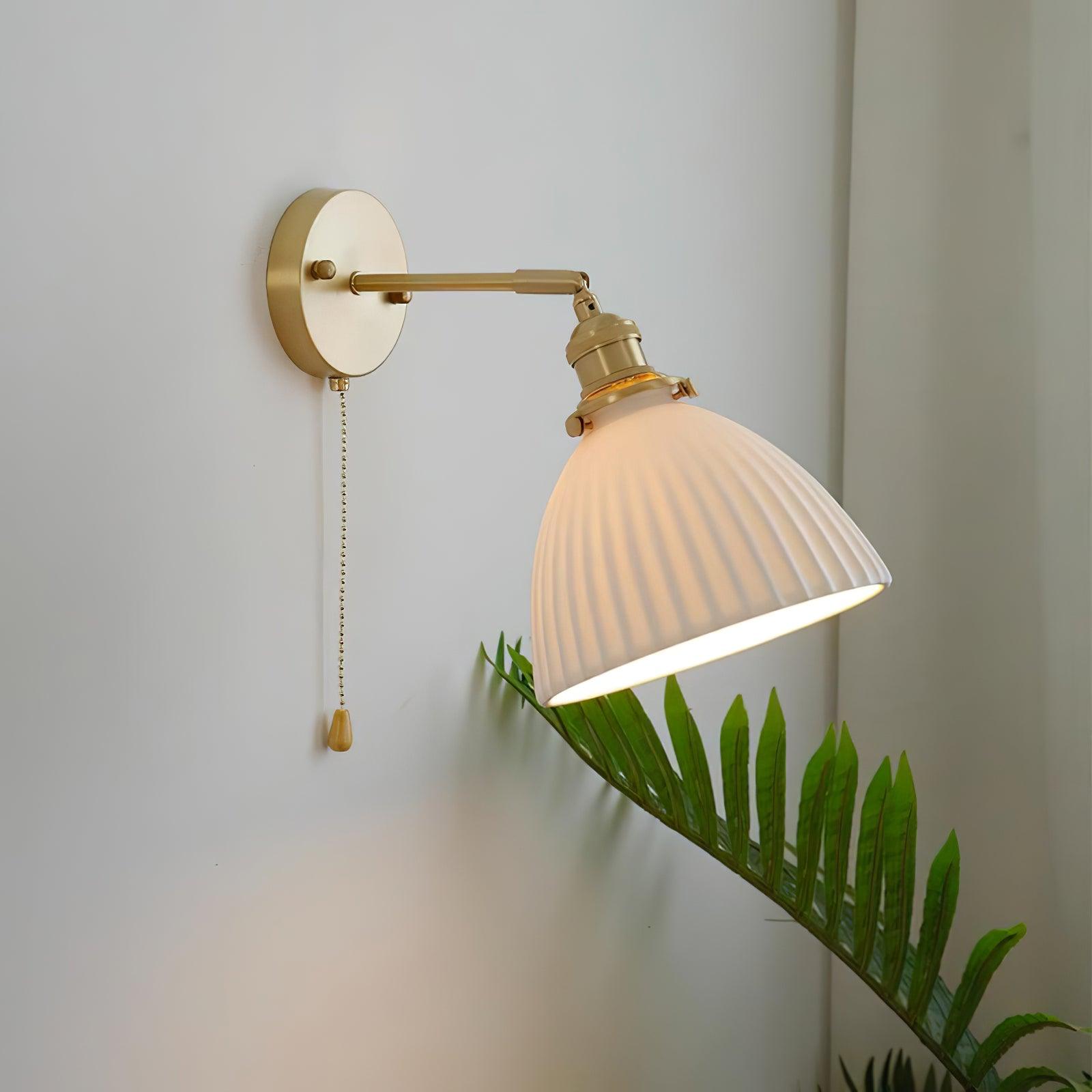 Angled lit white pleated shade on a brass arm above green fronds, with a round wall plate and teardrop-ended chain