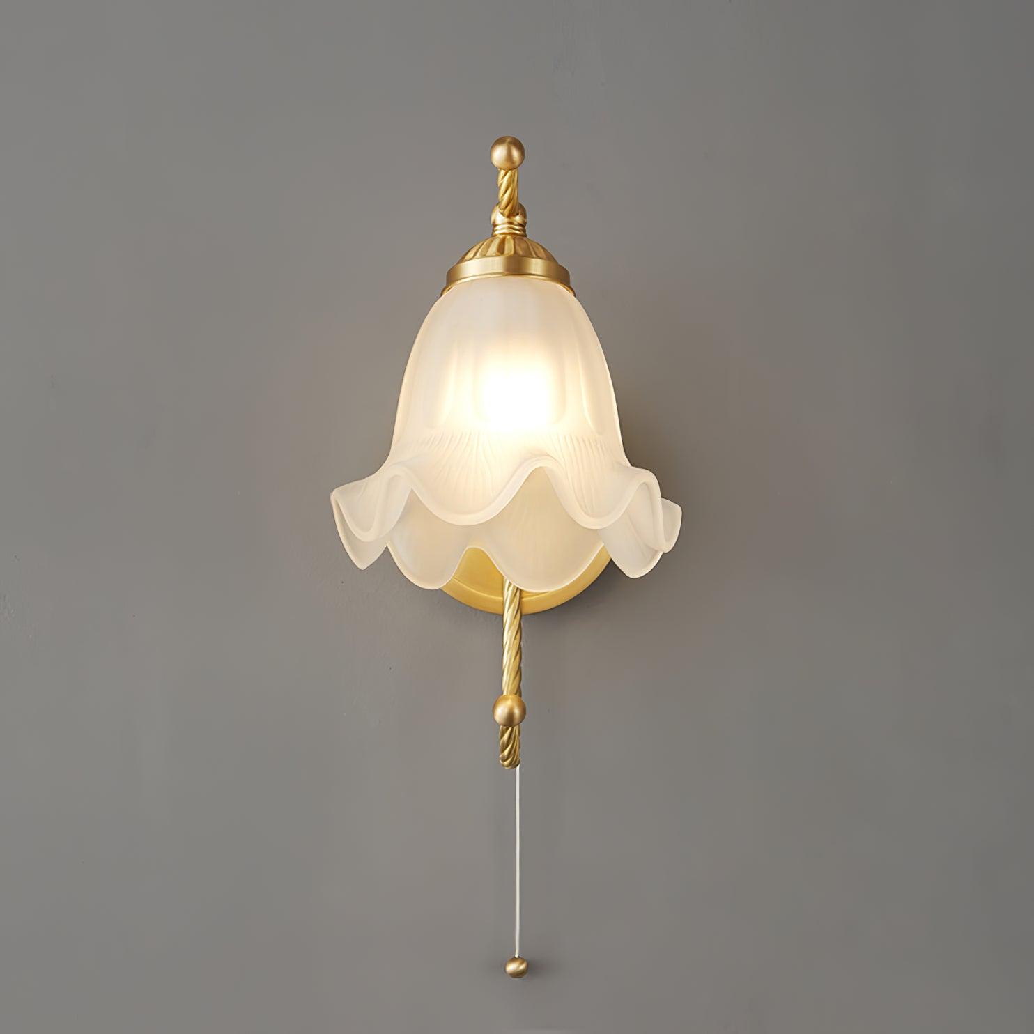 Front view of lit white scalloped glass wall lamp on grey, with twisted brass-finish arm above and below and pull cord beneath