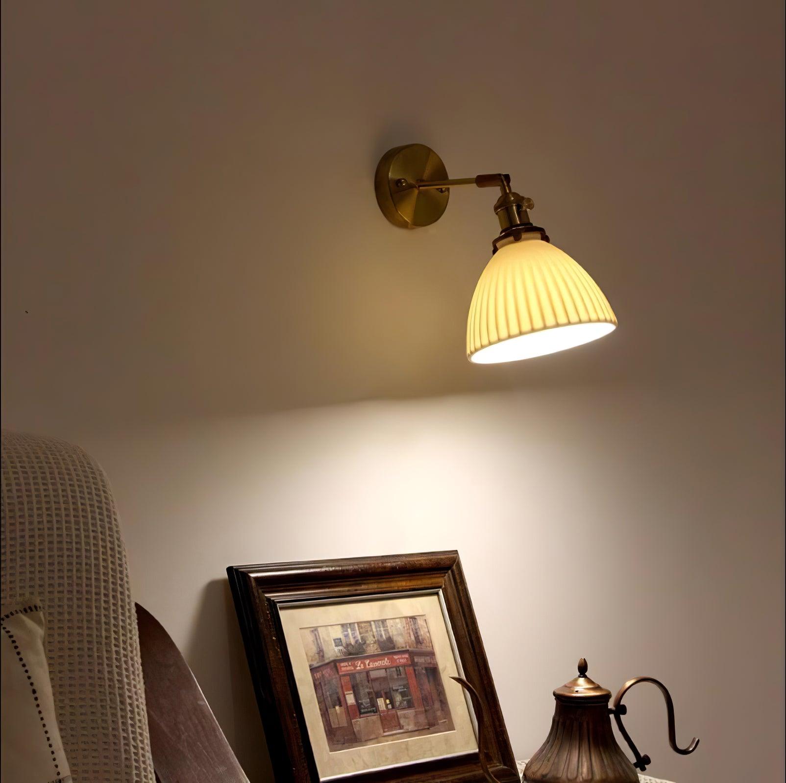 Warmly lit white pleated wall lamp above a framed picture and decorative metal pot on a lace-covered surface