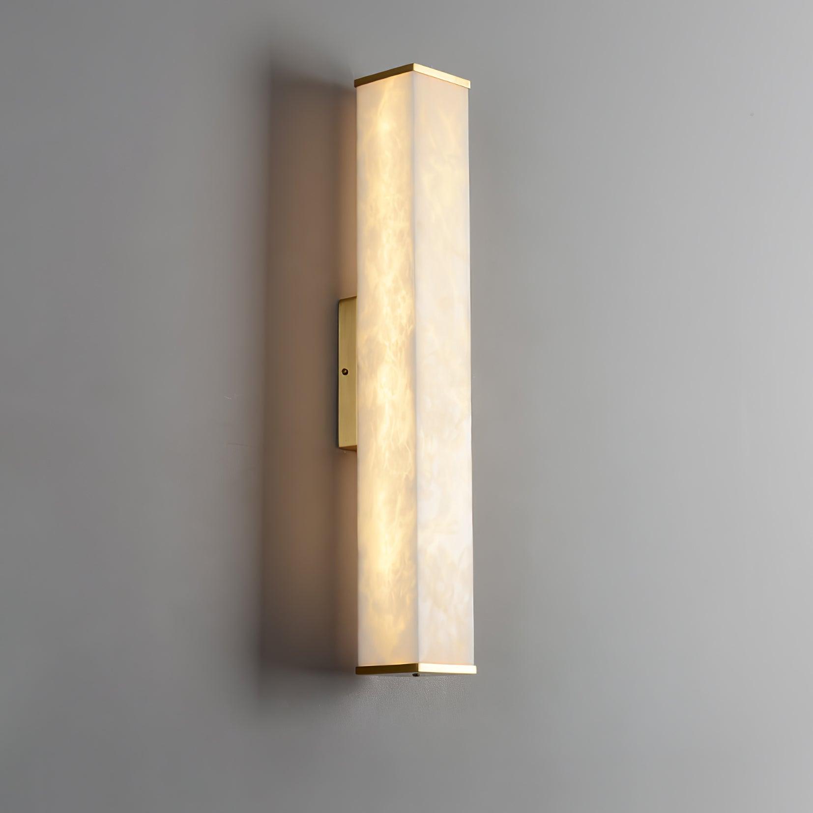Lit alabaster cuboid wall light with cloudy veining, brass end trims and a small mounting plate on a gray wall
