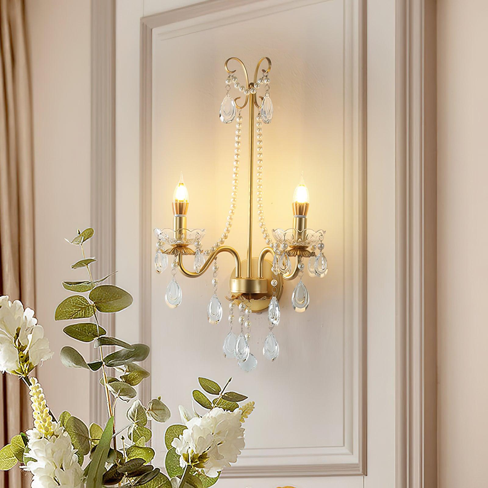Lit gold-and-clear wall light on pale moulded panel above white flowers and green round leaves, beside a curtain