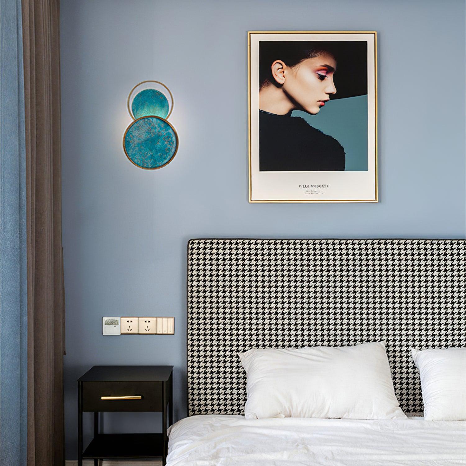 Single turquoise wall light on a blue bedroom wall beside a houndstooth headboard, black cabinet and framed portrait.