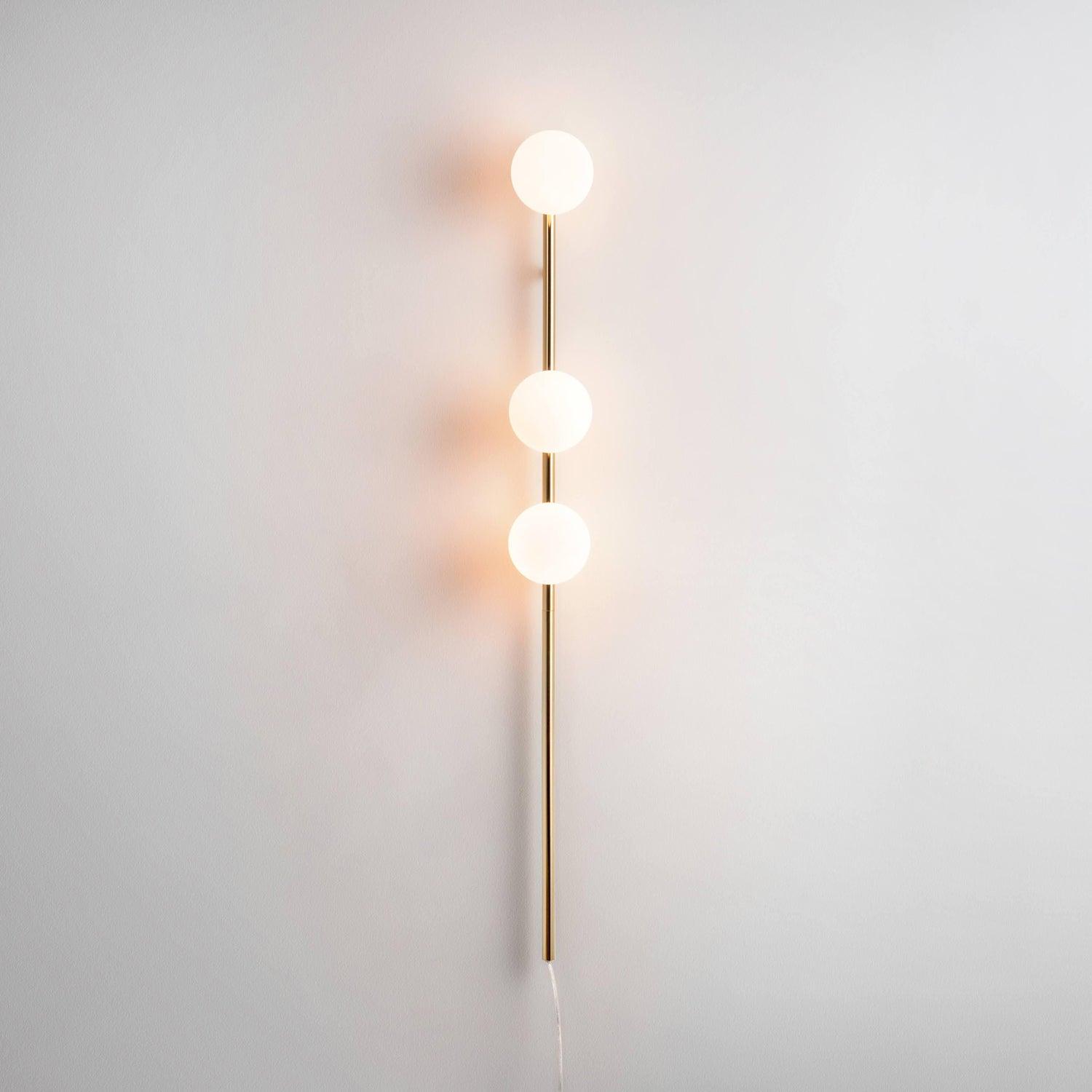 Gold three-globe wall light illuminated on pale wall, warm halos around white glass globes and long lower stem