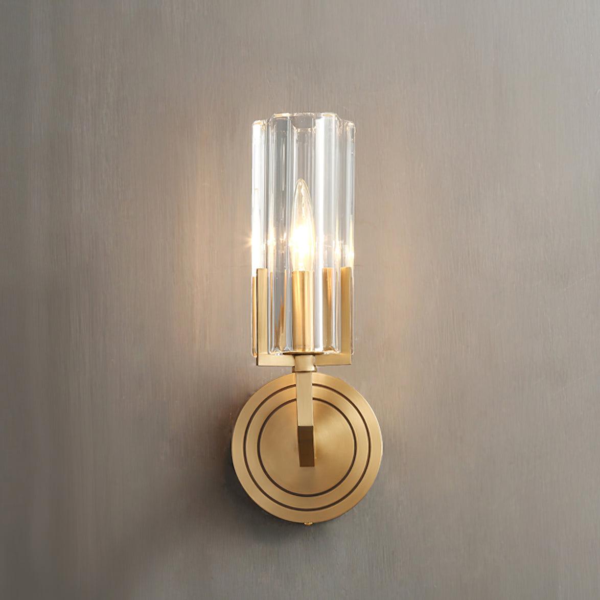 Frontal lit single fluted glass shade above ringed circular brass plate on grey wall, with narrow arm seen head-on