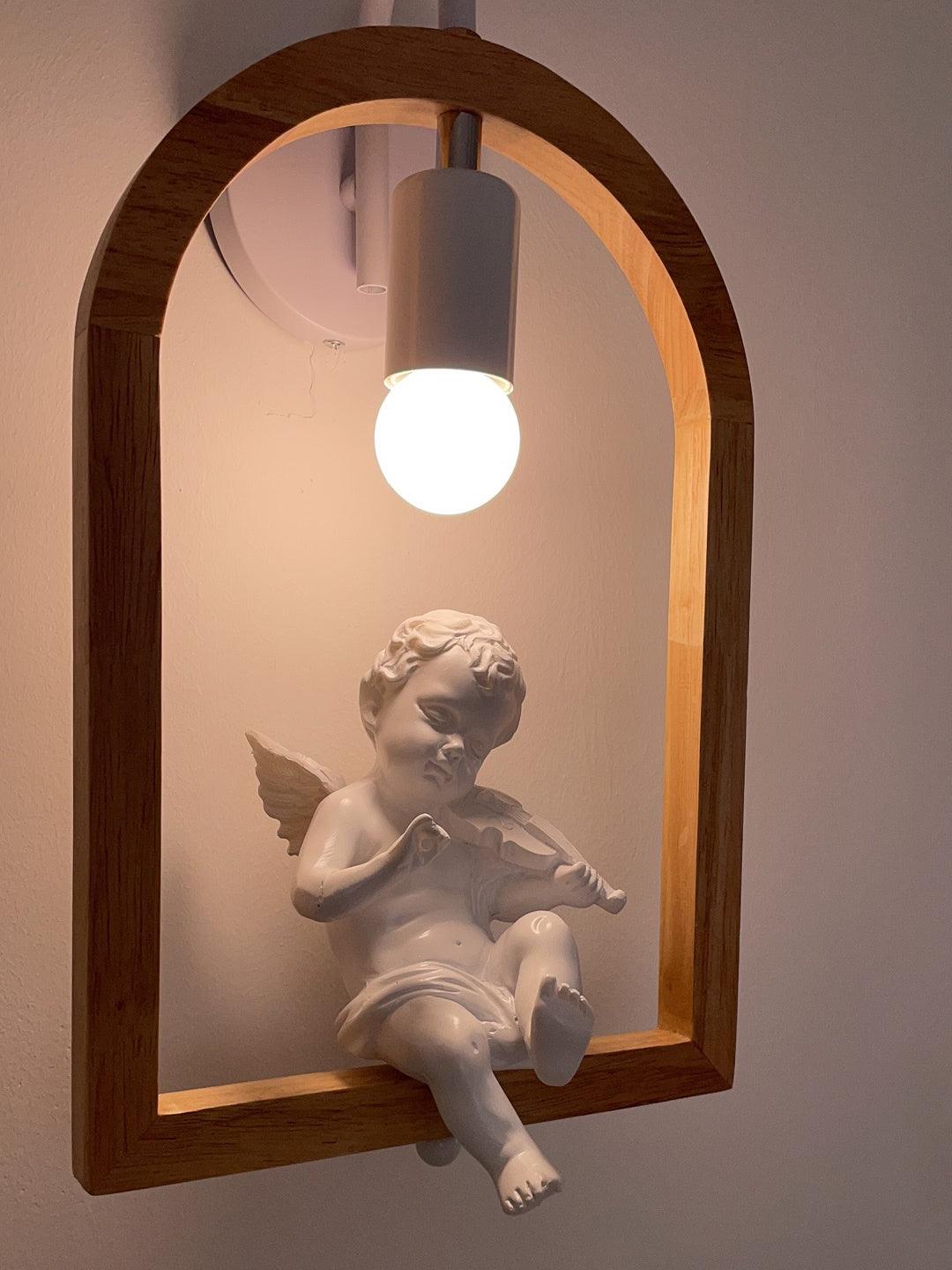 Angled close-up of the white cherub playing violin, with wings and legs around the lower wooden rail beneath a lit bulb