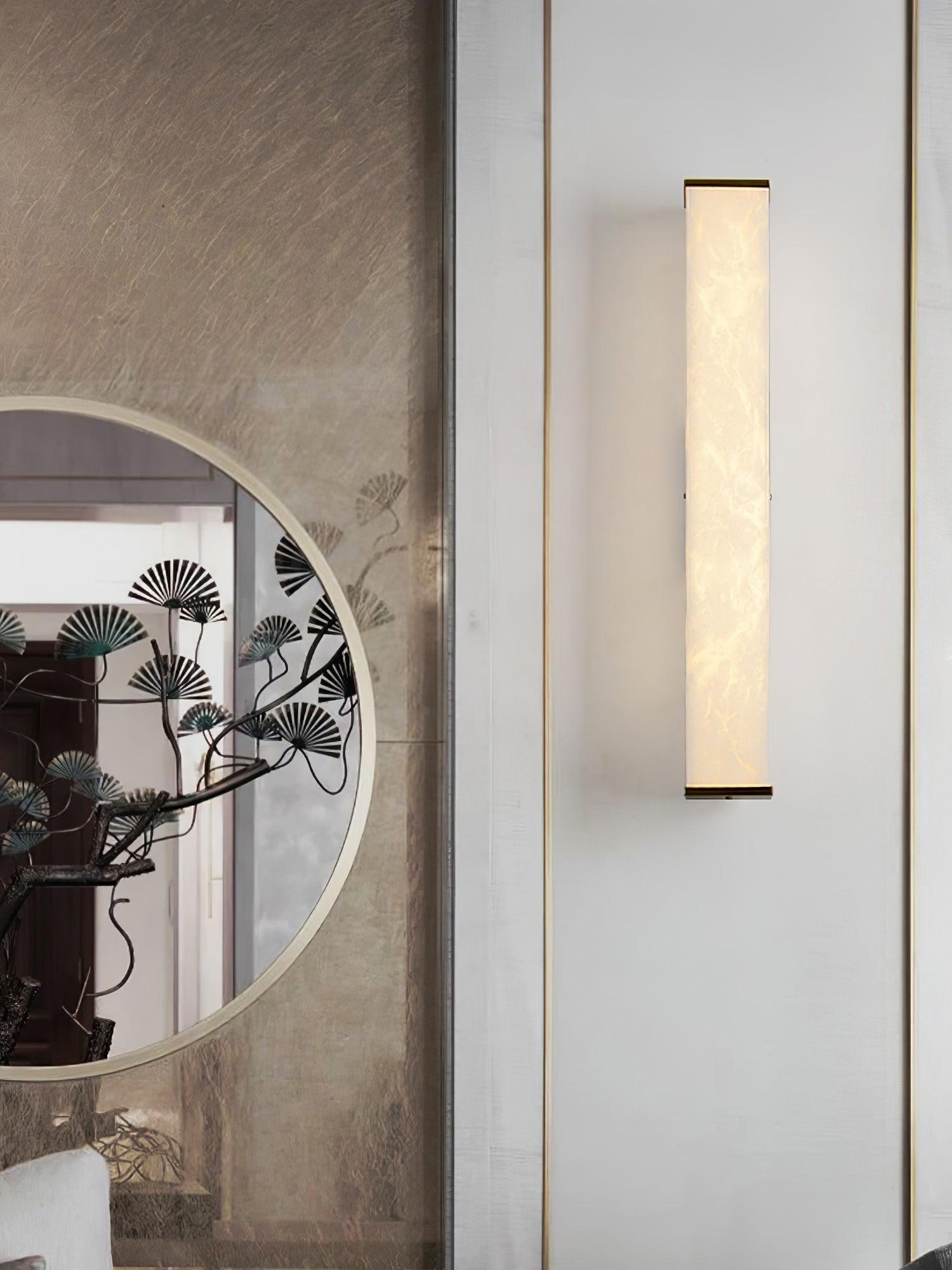 Front view of the glowing stone wall light with narrow brass end bands beside a round mirror on a pale paneled wall