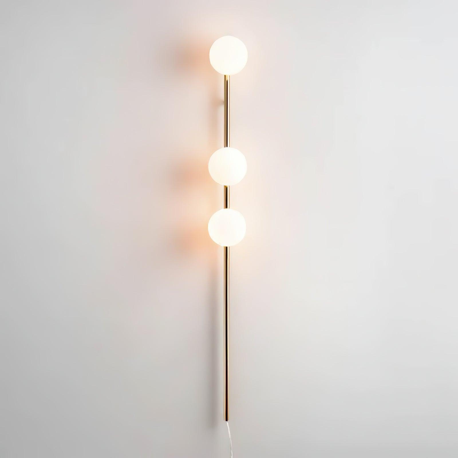 Gold vertical wall light illuminated on pale wall, three glowing glass globes, reflective stem and pale cable