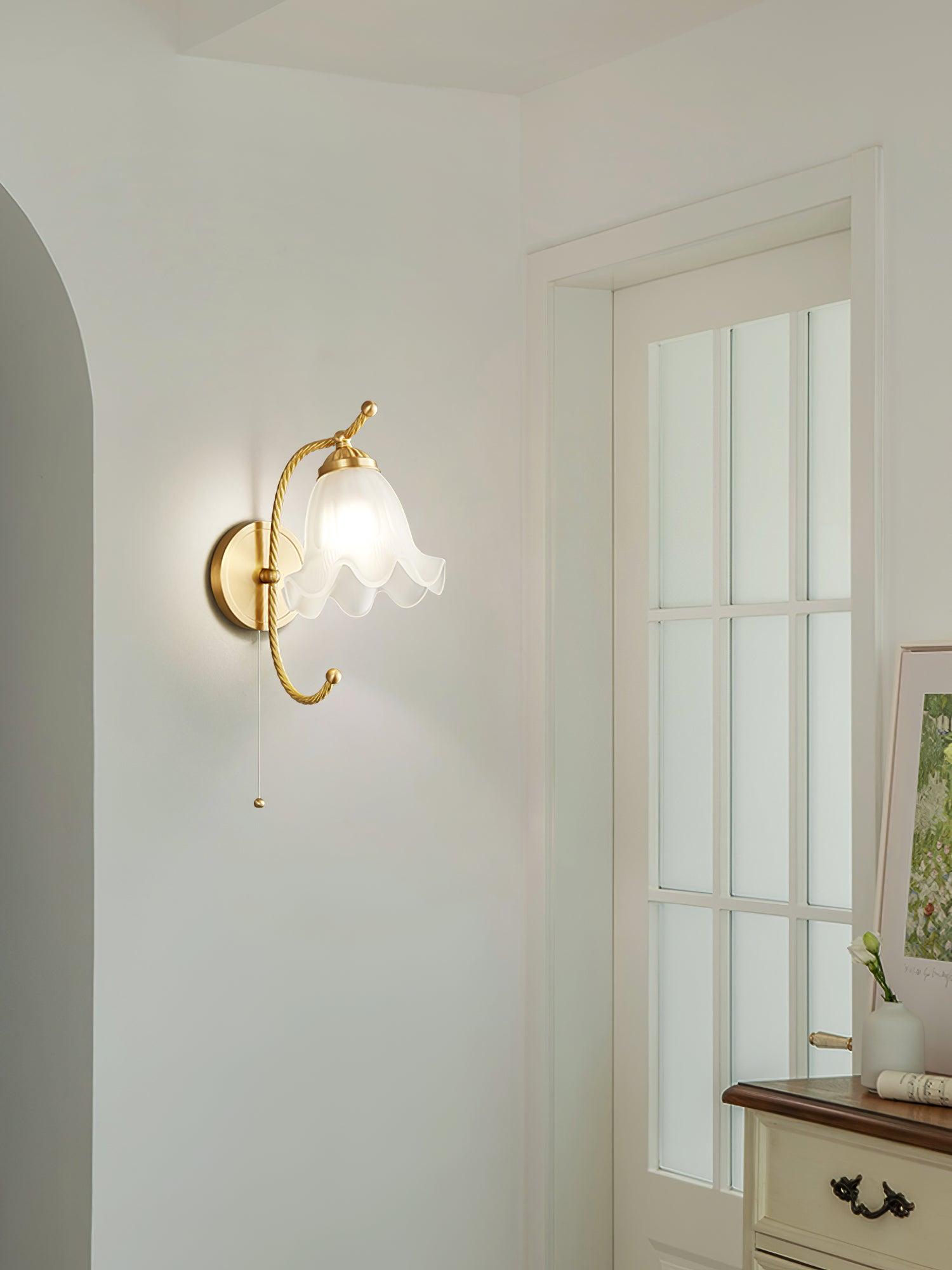 Lit brass gooseneck wall lamp beside glass-paned door and dresser with framed floral artwork, showing pull cord at left