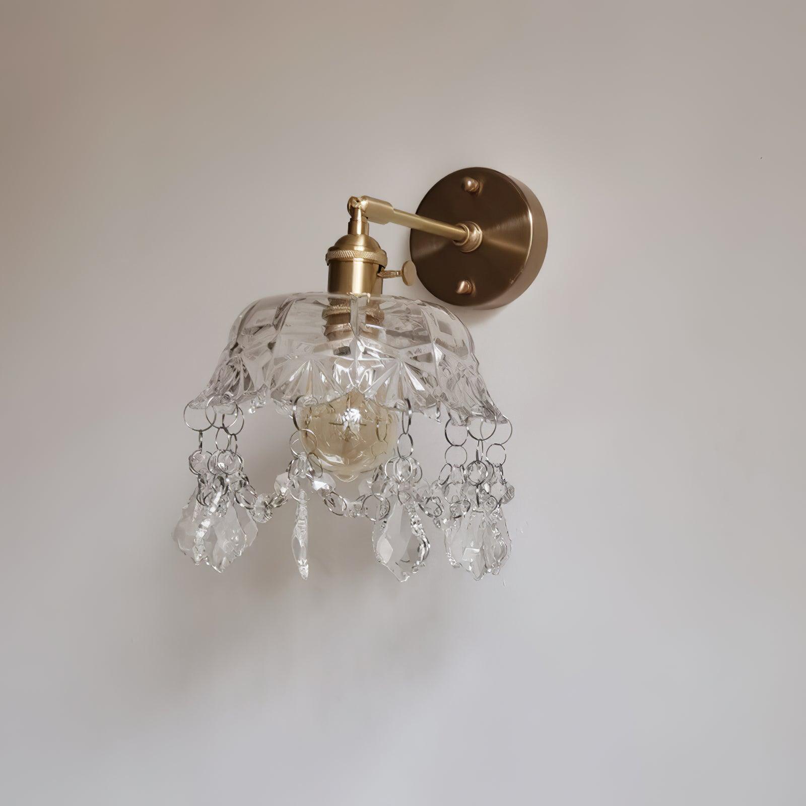 Unlit clear bell and crystal drop fringe below a gold-tone arm and round wall plate on a pale wall