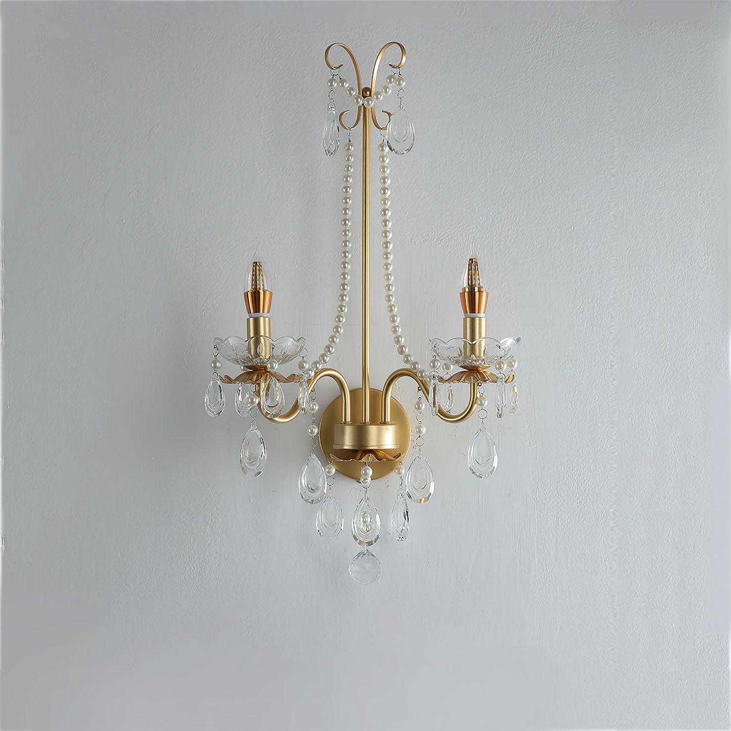 Unlit front gold-finish fixture on textured white wall with two candle bulbs, clear scalloped bowls, pale beads and teardrop-shaped drops